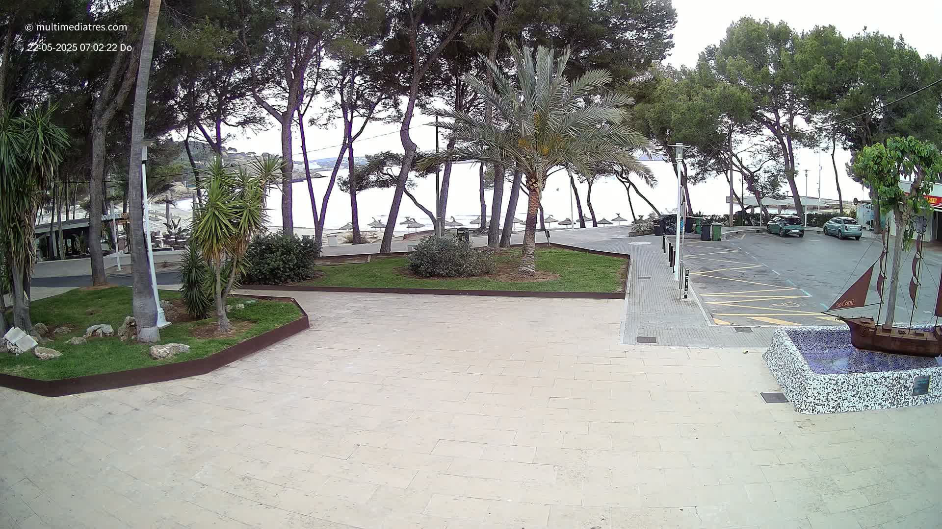 A paved area with a small park containing palm trees and other vegetation overlooks a calm beach with several umbrellas, all under an overcast sky.