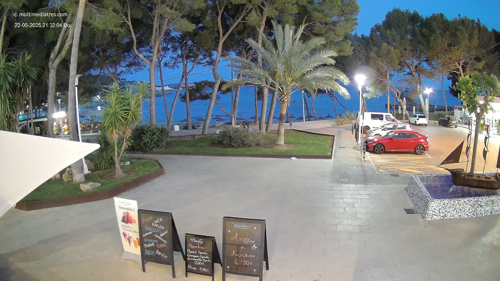 A seaside plaza at dusk, featuring palm trees, parked cars, and restaurant signage, with a calm blue sea visible in the background.