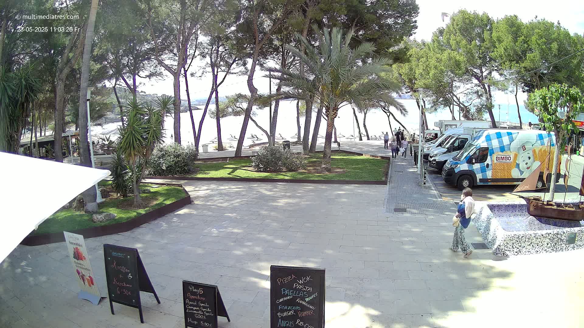 A paved plaza with several food stands, lined with pine and palm trees, overlooks a sunny beach with people and parked vans.