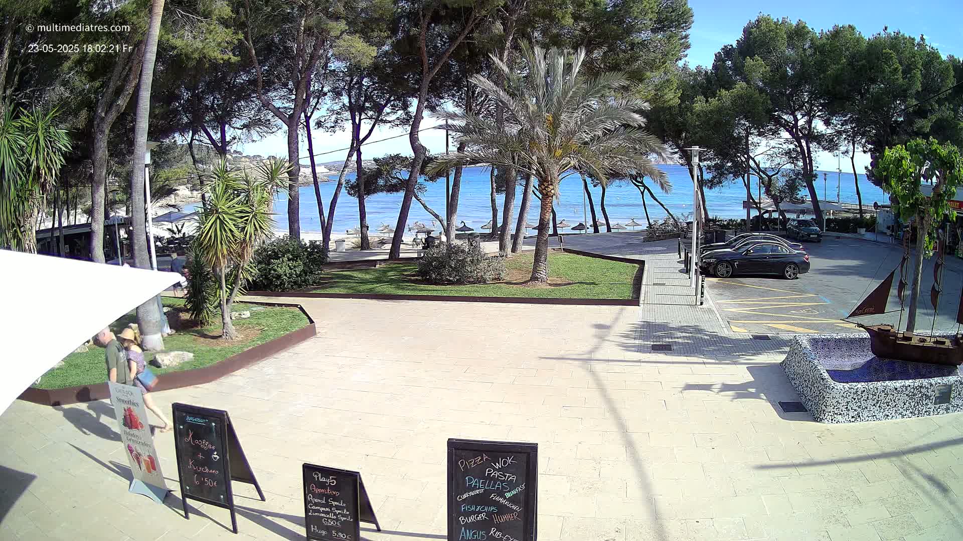 A paved plaza with palm trees and other vegetation overlooks a sandy beach and calm, blue ocean under a sunny sky.