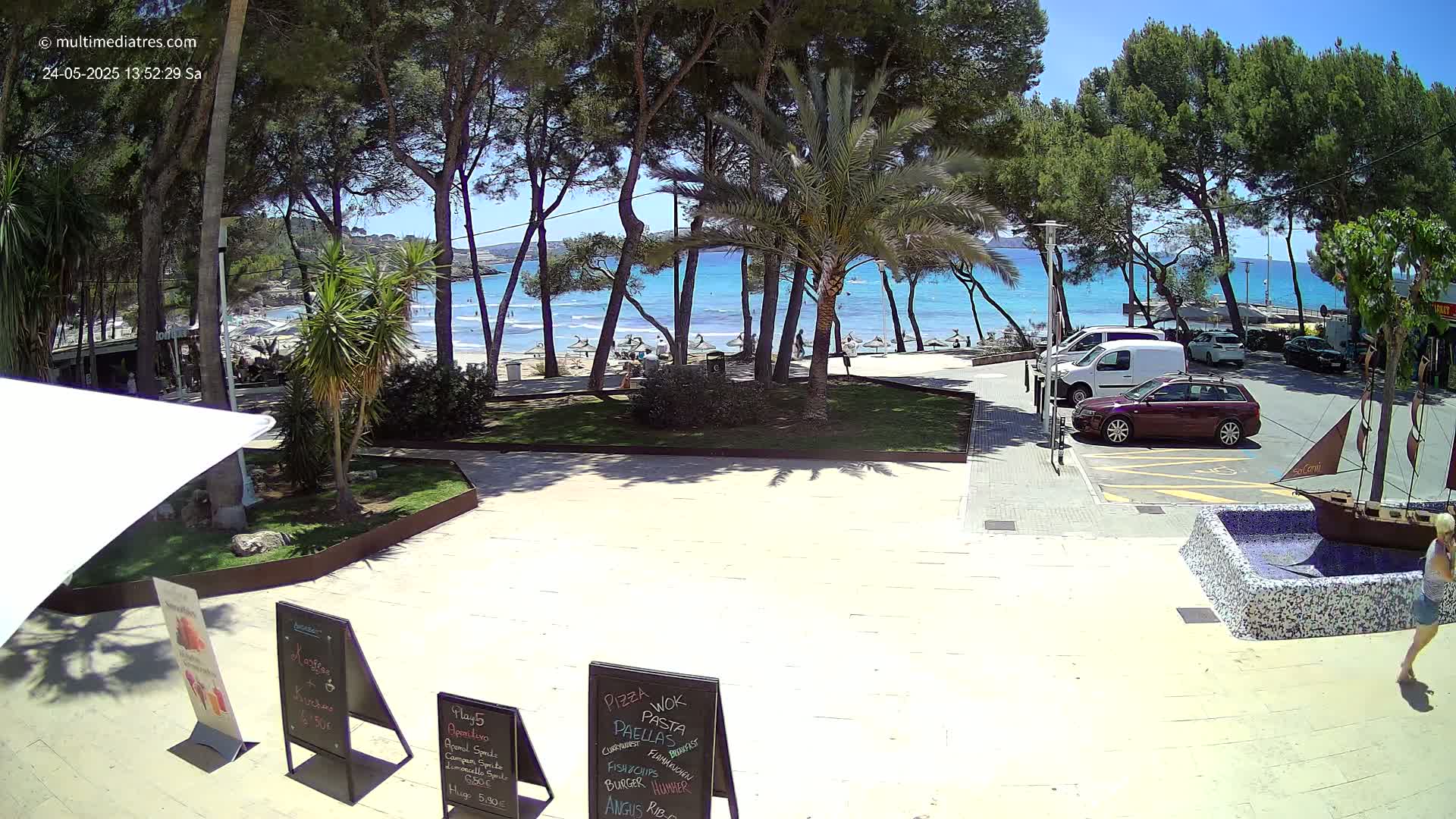 A sunny beach scene is visible through a grove of trees, with parked cars and a small decorative boat fountain in the foreground.