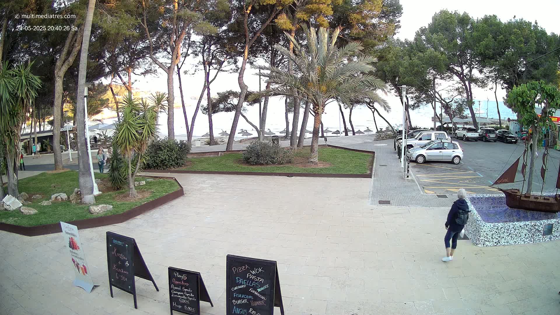 A paved plaza with palm trees and grassy areas overlooks a calm ocean beach with beach umbrellas under a partly cloudy sky.