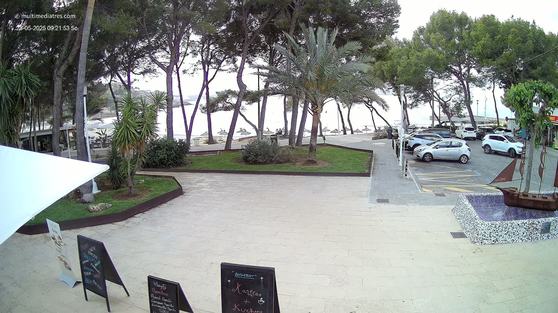 A paved plaza with a small park and several cars parked nearby overlooks a beach with calm water under an overcast sky.