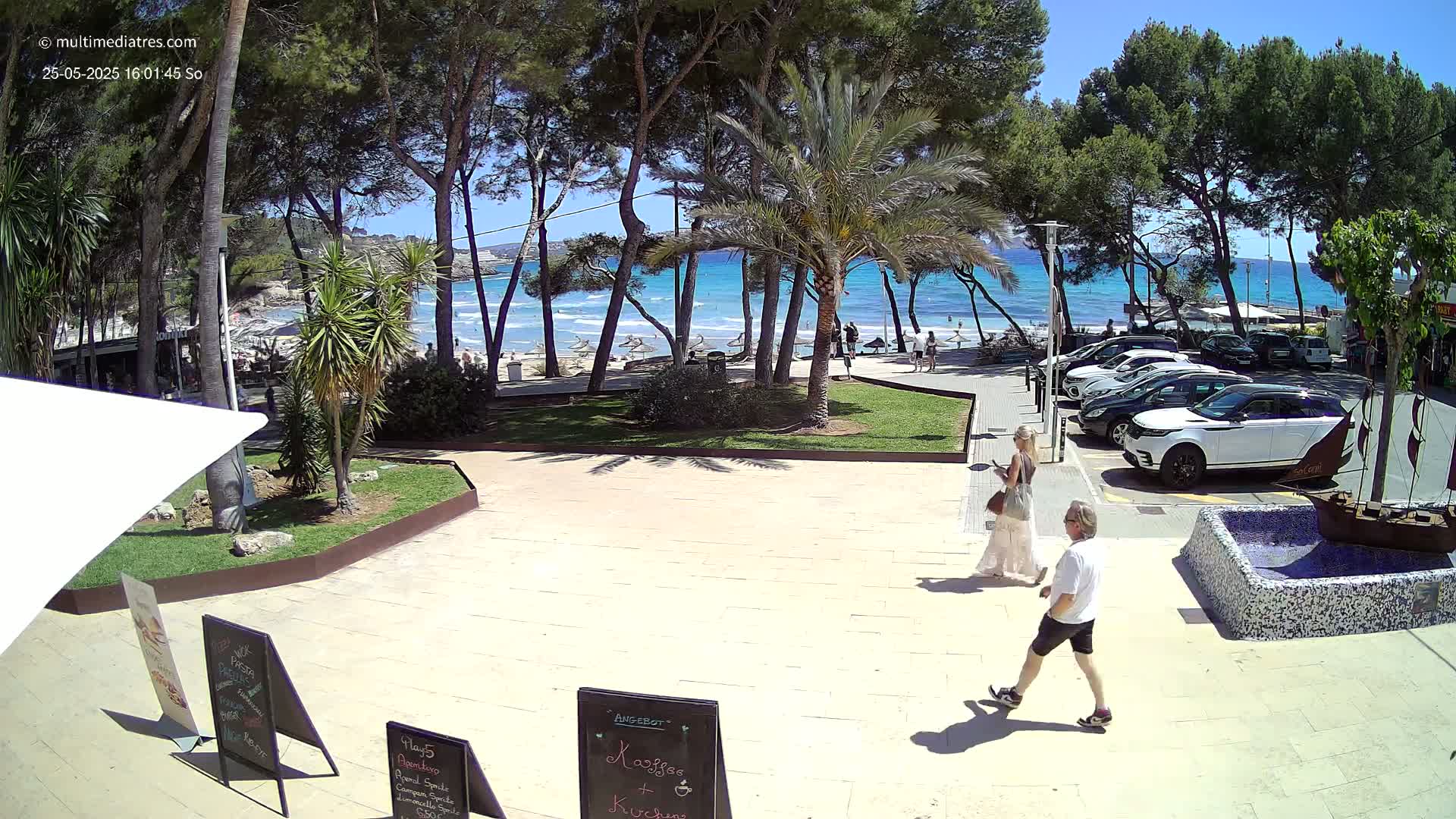 A paved area with two people walking, near a beach with turquoise water under a sunny sky, surrounded by trees and parked cars.