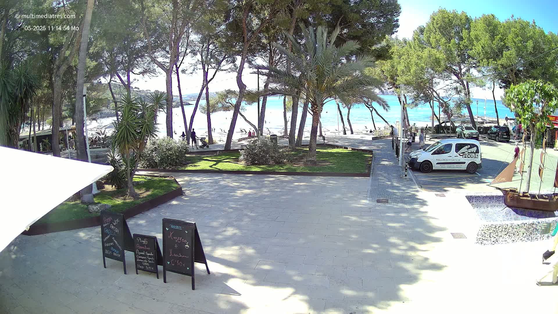 A paved area with beachside menus leads to a sandy beach and turquoise water, viewed through a grove of pine and palm trees under a sunny sky.