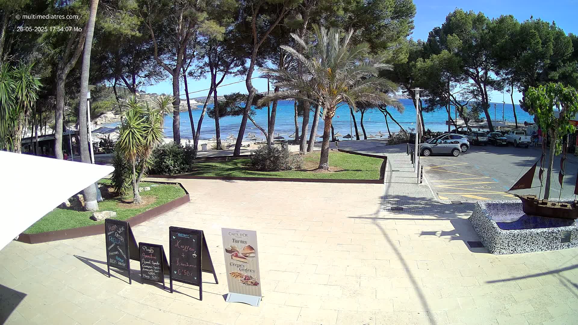 A sunny beachfront plaza scene with palm trees, a parking lot, and a small decorative ship fountain.