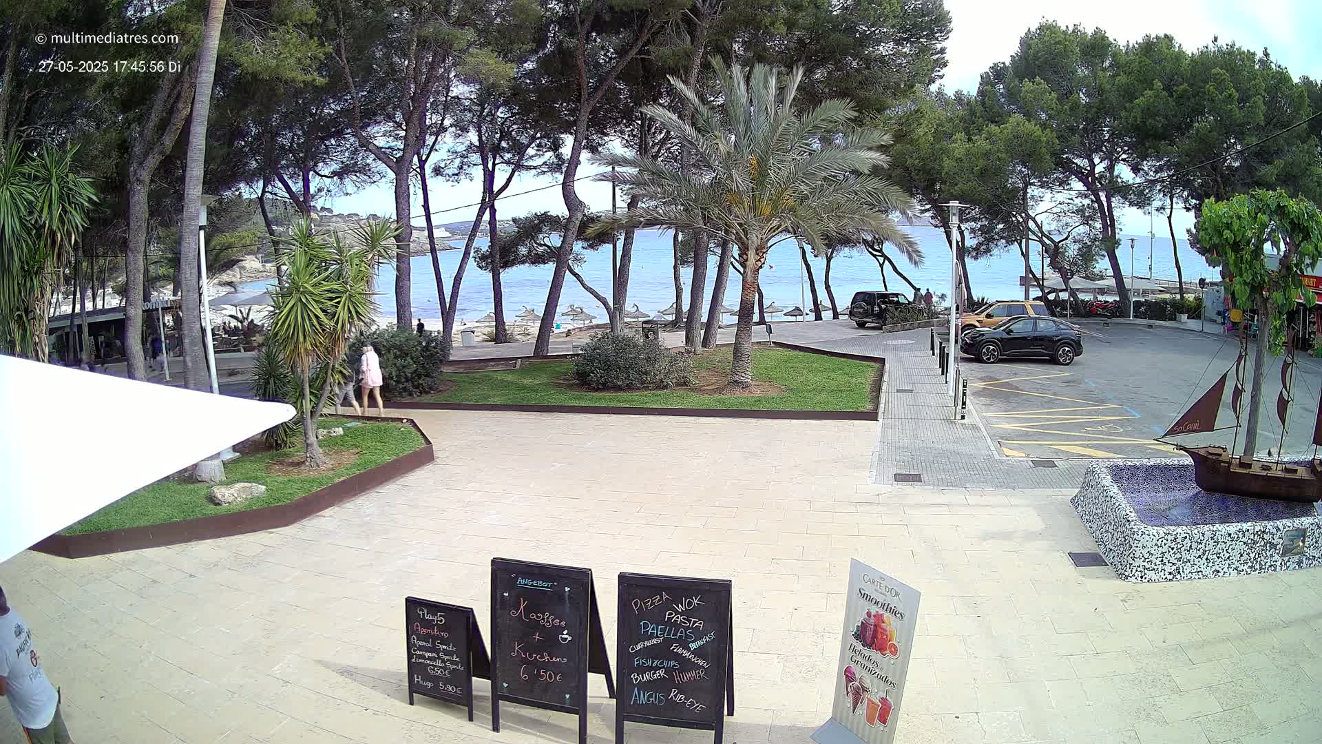 A partly sunny beach scene is visible through a line of trees from a plaza with several cars parked, some people walking, and several menu boards on the ground.