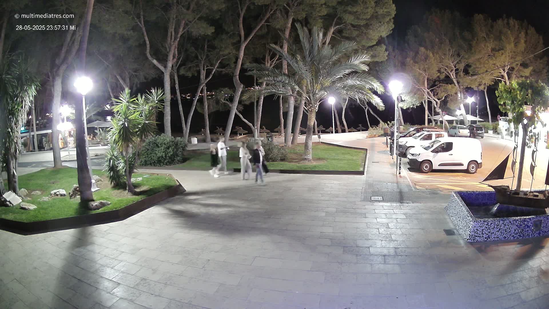 A paved plaza at night, illuminated by streetlights, shows several people walking among palm trees and other vegetation, with a parking lot visible in the background.