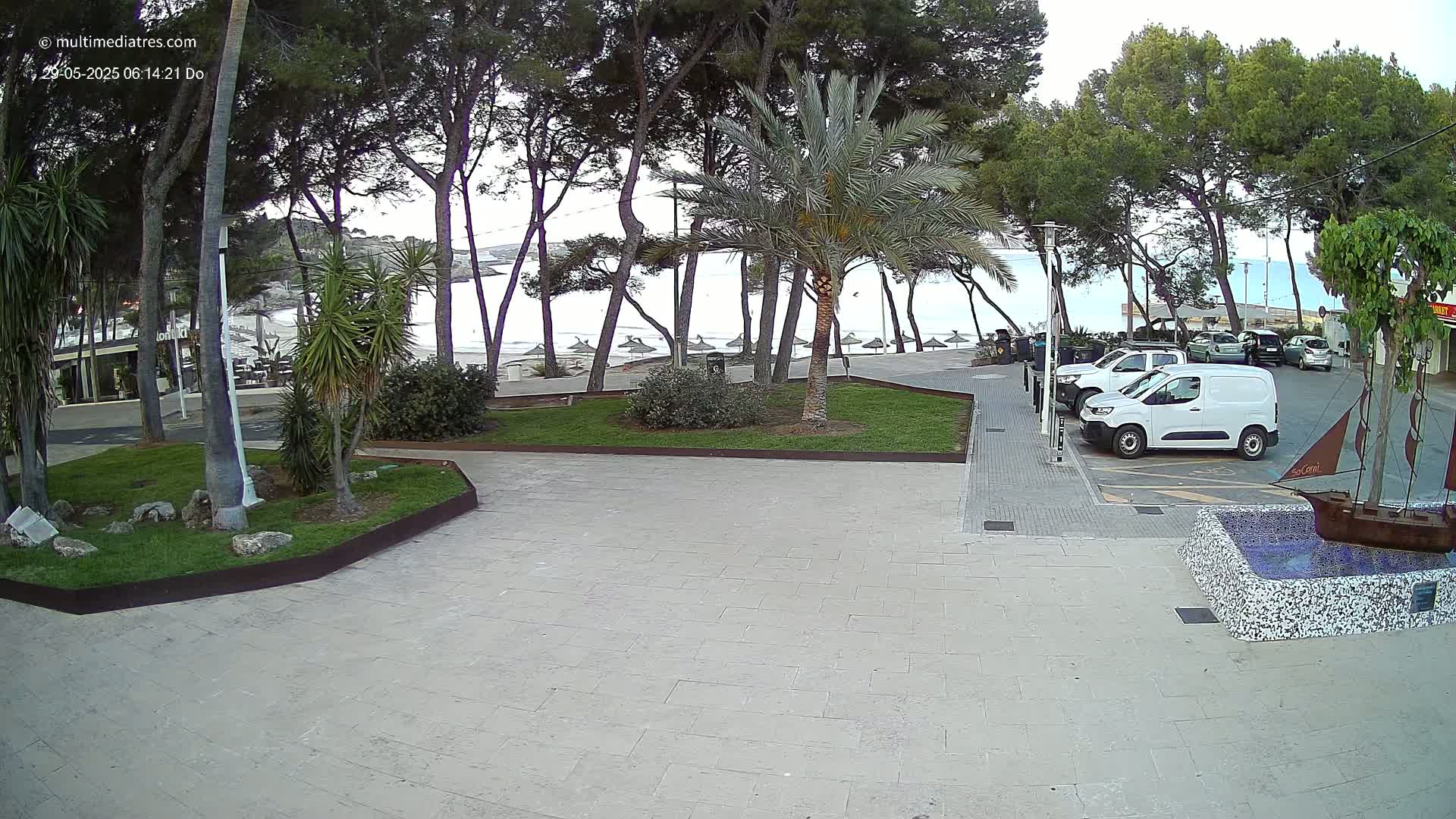 A paved plaza with a grassy area and several trees overlooks a calm beach with beach umbrellas under a mostly clear sky.