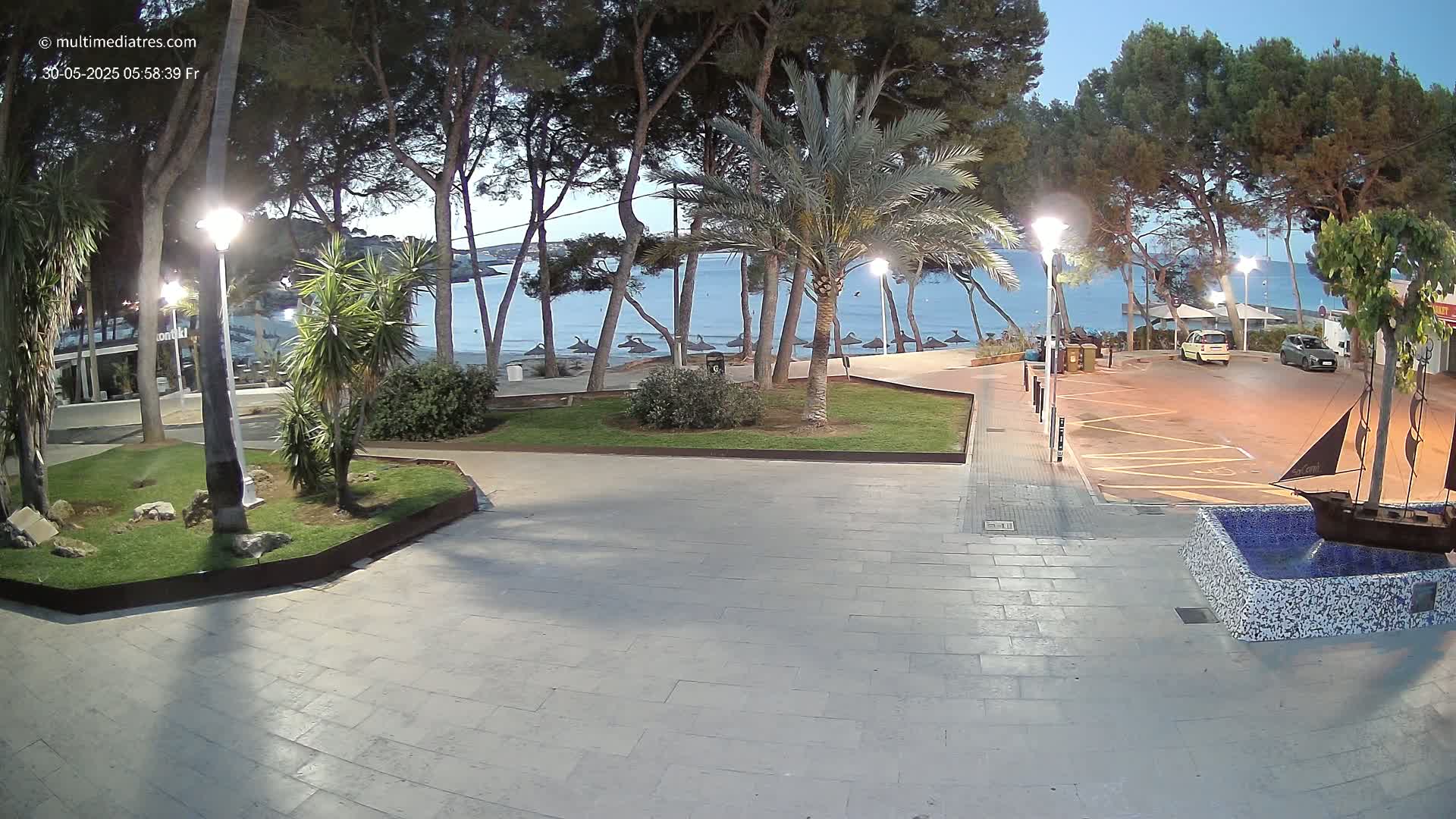 A paved plaza at dusk overlooks a calm beach with closed umbrellas, lined with palm trees and pines under nighttime lighting.