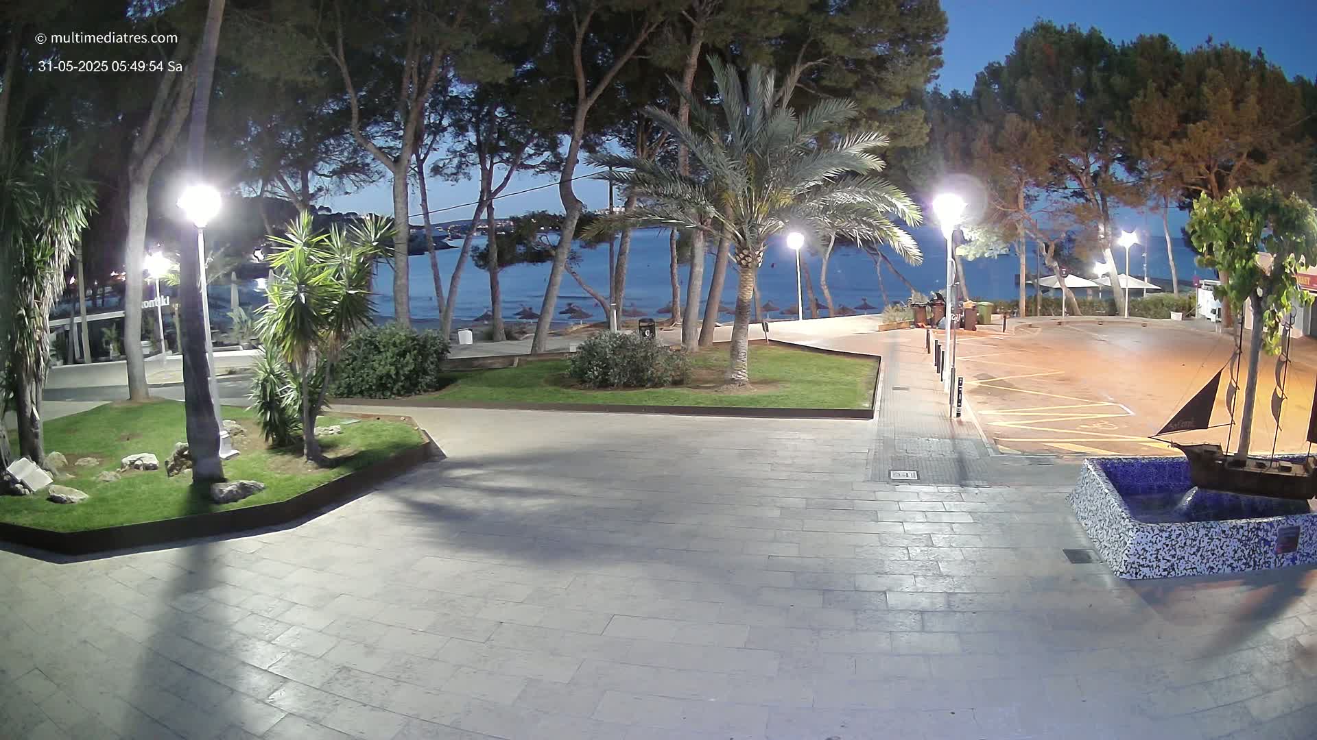 A paved plaza at night, illuminated by lamps, overlooks a calm beach with palm trees and a tranquil sea under a twilight sky.