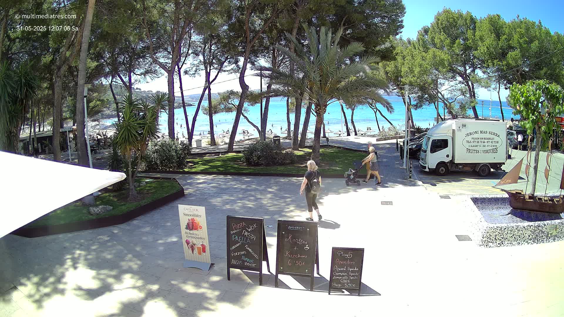 A sunny beach scene is viewed from a paved area through a grove of pine trees, where people stroll near outdoor cafe menu boards and a food truck.