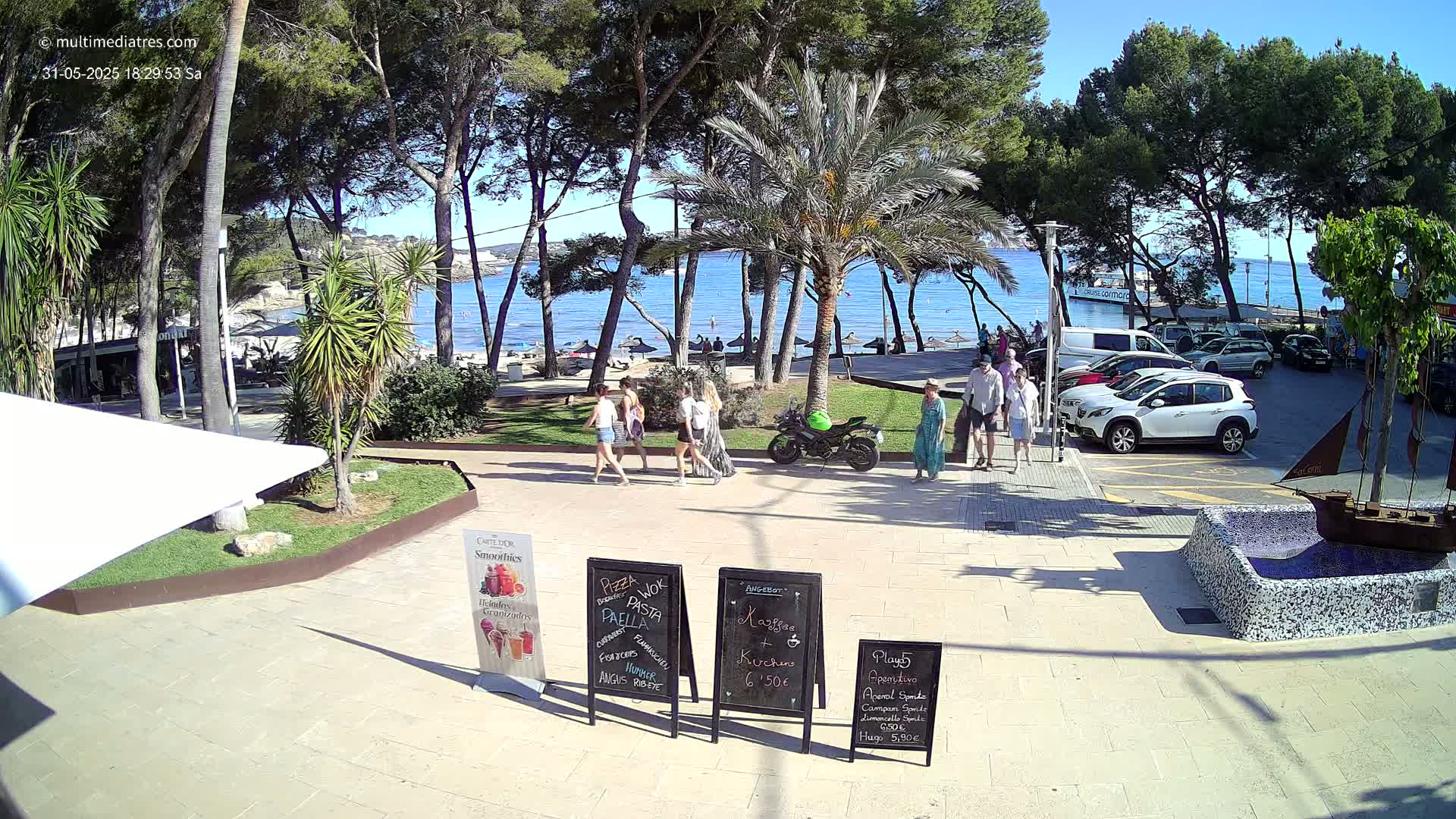 A sunny beach scene is visible through a grove of palm and pine trees, with people walking along a paved area near a parking lot and a small beach with umbrellas.