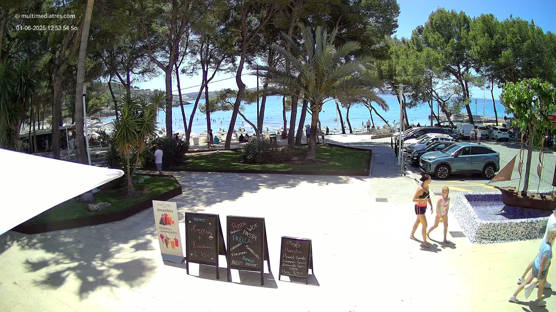A sunny beach scene with people relaxing on the sand and others walking along a paved area near a parking lot, under a canopy of pine trees.
