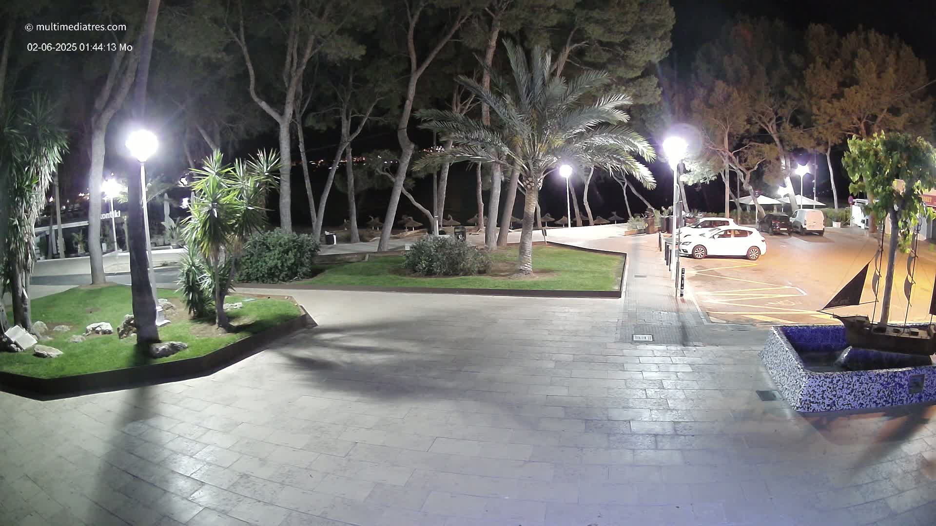 A nighttime view of a paved plaza with palm trees and other landscaping, illuminated by lampposts, next to a parking lot containing several cars.