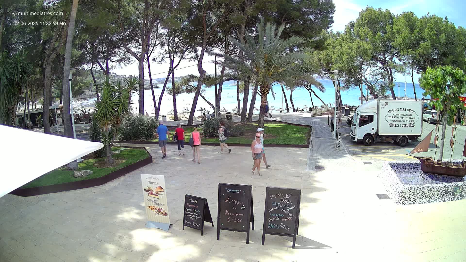 A sunny day at a beachside plaza shows several people walking near palm trees and food stands, with a view of a sandy beach and turquoise water beyond.