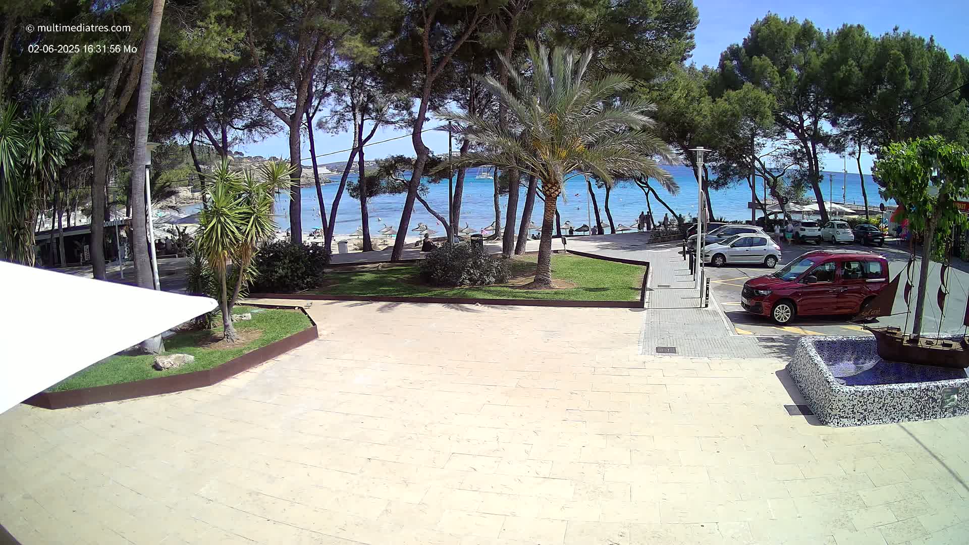 A paved plaza overlooks a sunny beach with clear blue water, surrounded by palm trees and pine trees.