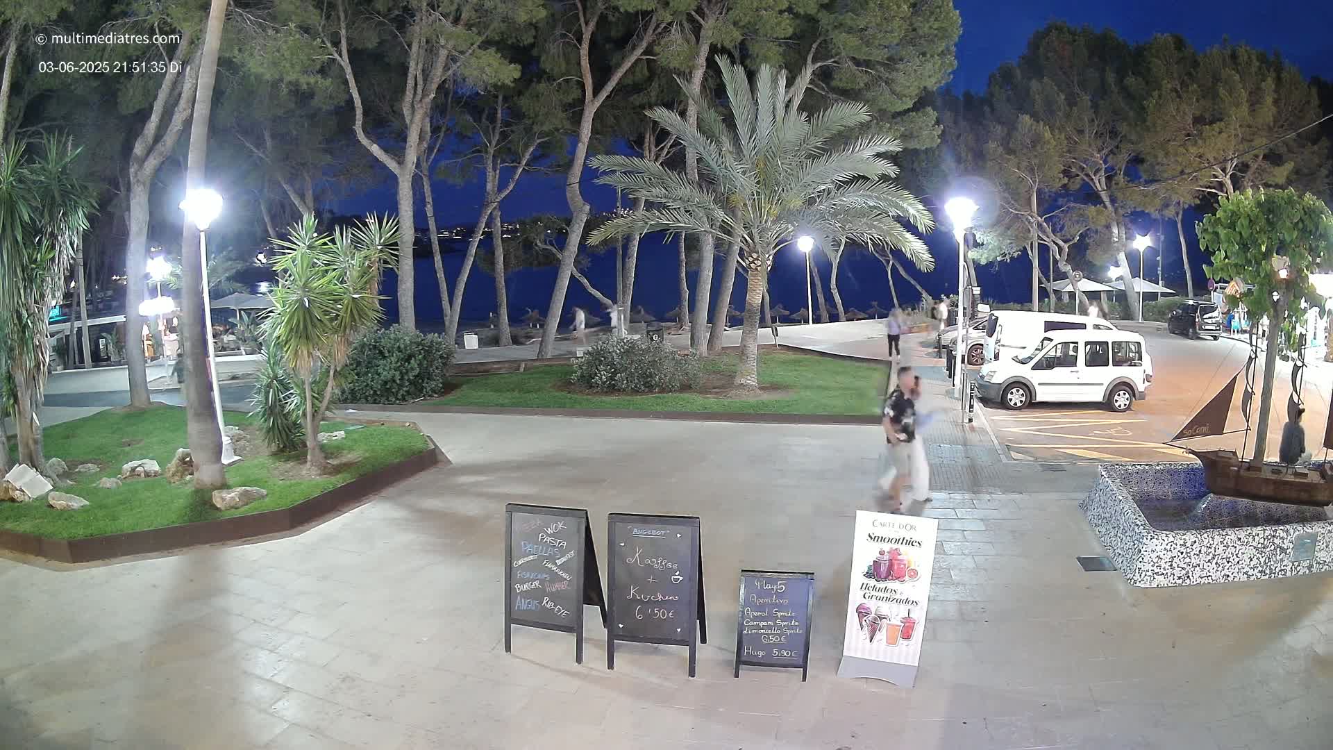 A nighttime scene shows a paved plaza with palm trees, people walking, and a white van parked near a restaurant with menu boards.