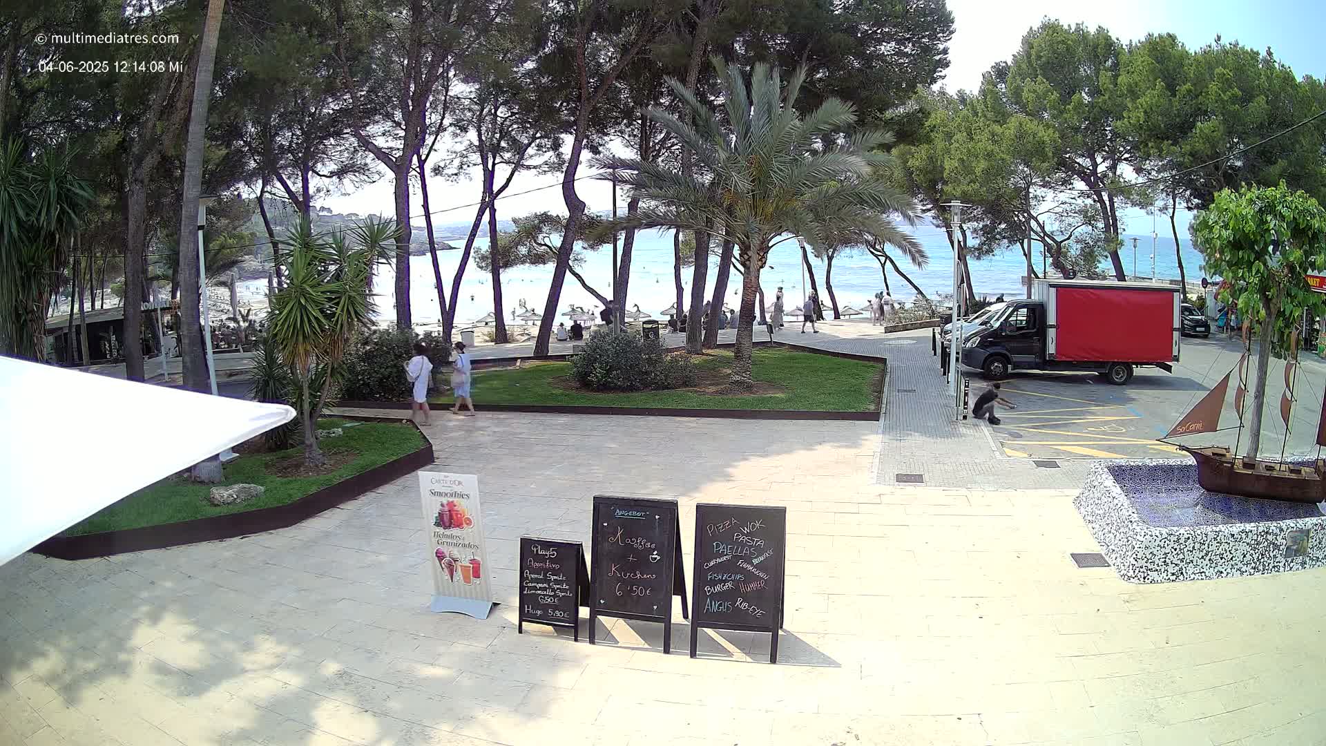 A sunny beachfront plaza features palm trees, a sandy beach with people, and a red delivery truck parked nearby.