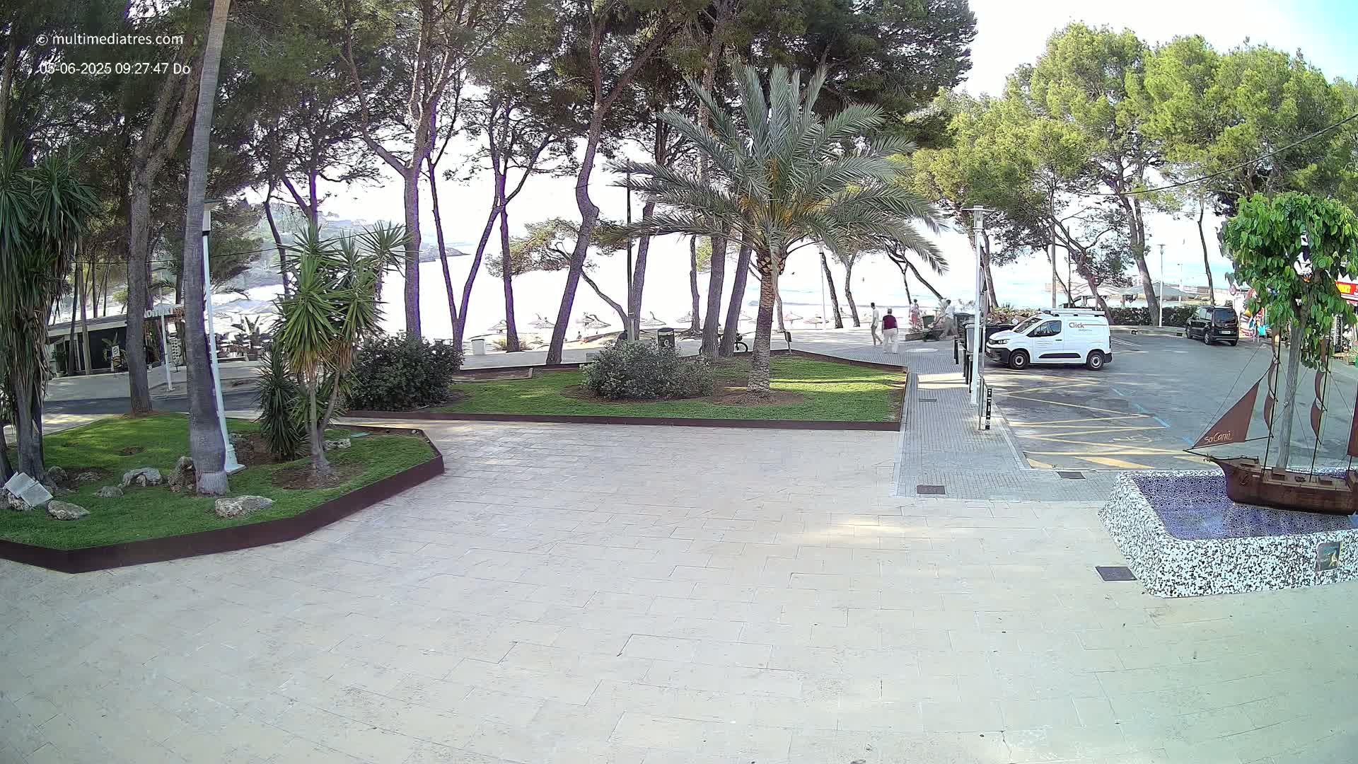 A paved plaza with a small grassy area and palm trees, overlooking a beach on a sunny day.