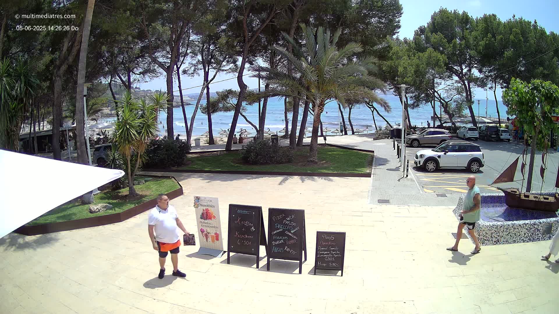 A sunny beach scene is visible from a beachfront patio area with several people walking around and parked cars nearby, under a mostly clear sky.