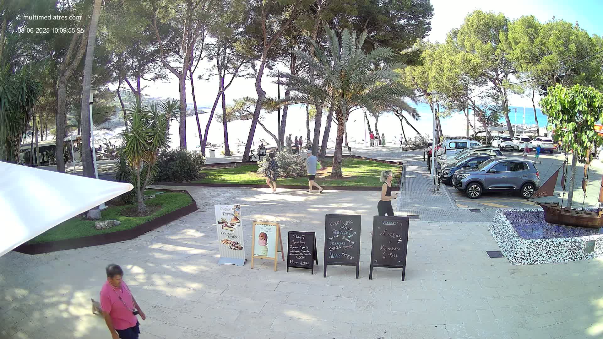 A sunny outdoor scene shows a paved area with several people walking, menus on easels, parked cars, and palm trees leading to a beach visible through the trees.