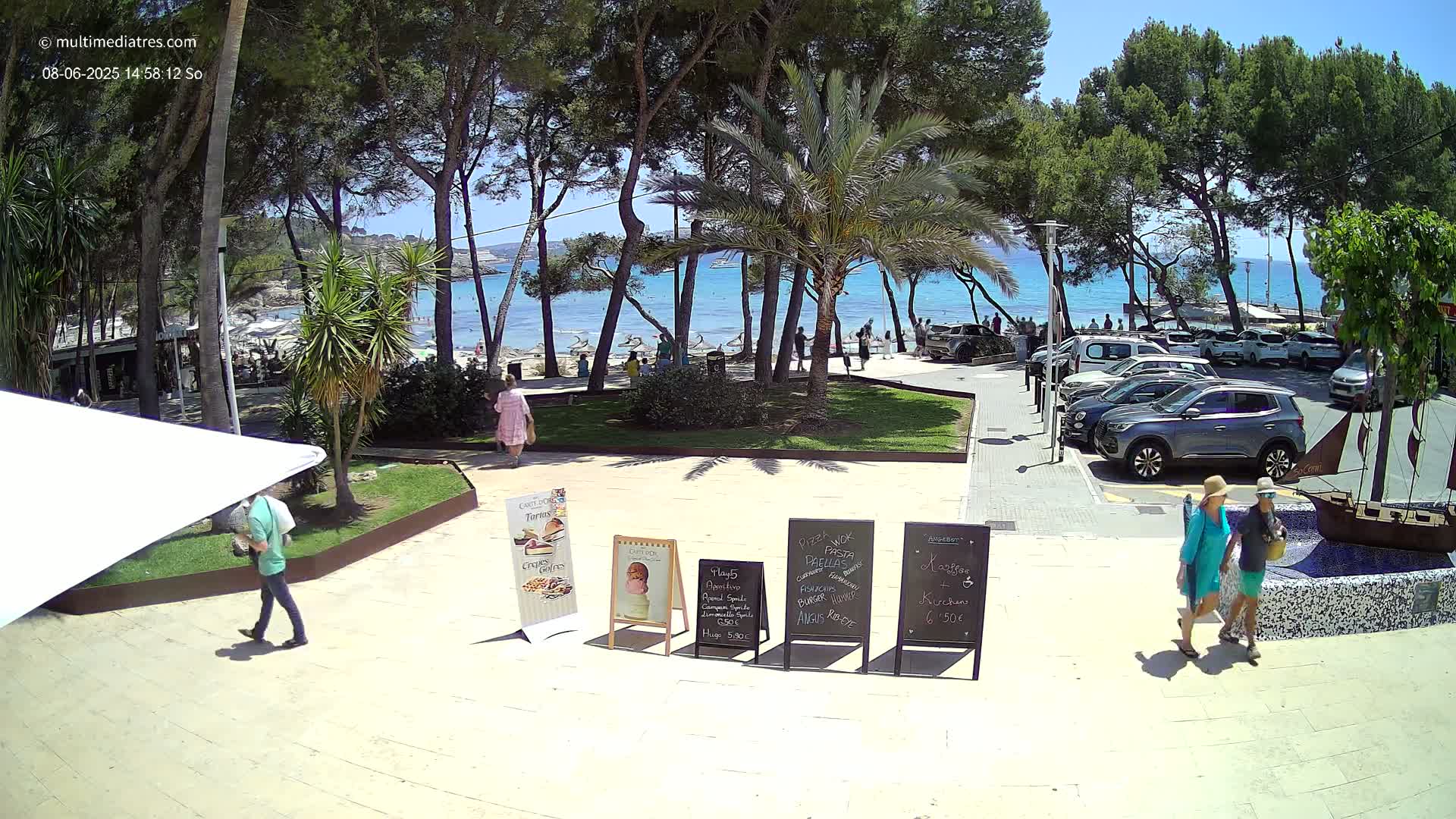 A sunny beach scene is visible through a grove of trees, with people relaxing on the beach and cars parked nearby; several people are walking on a paved area near outdoor restaurant menu boards.