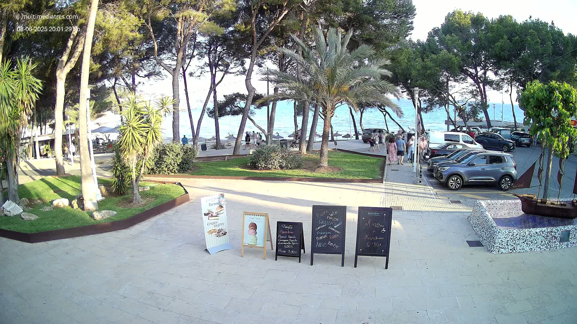A seaside plaza with palm trees and pine trees, under a clear evening sky, features several parked cars and a few people walking near a beach.