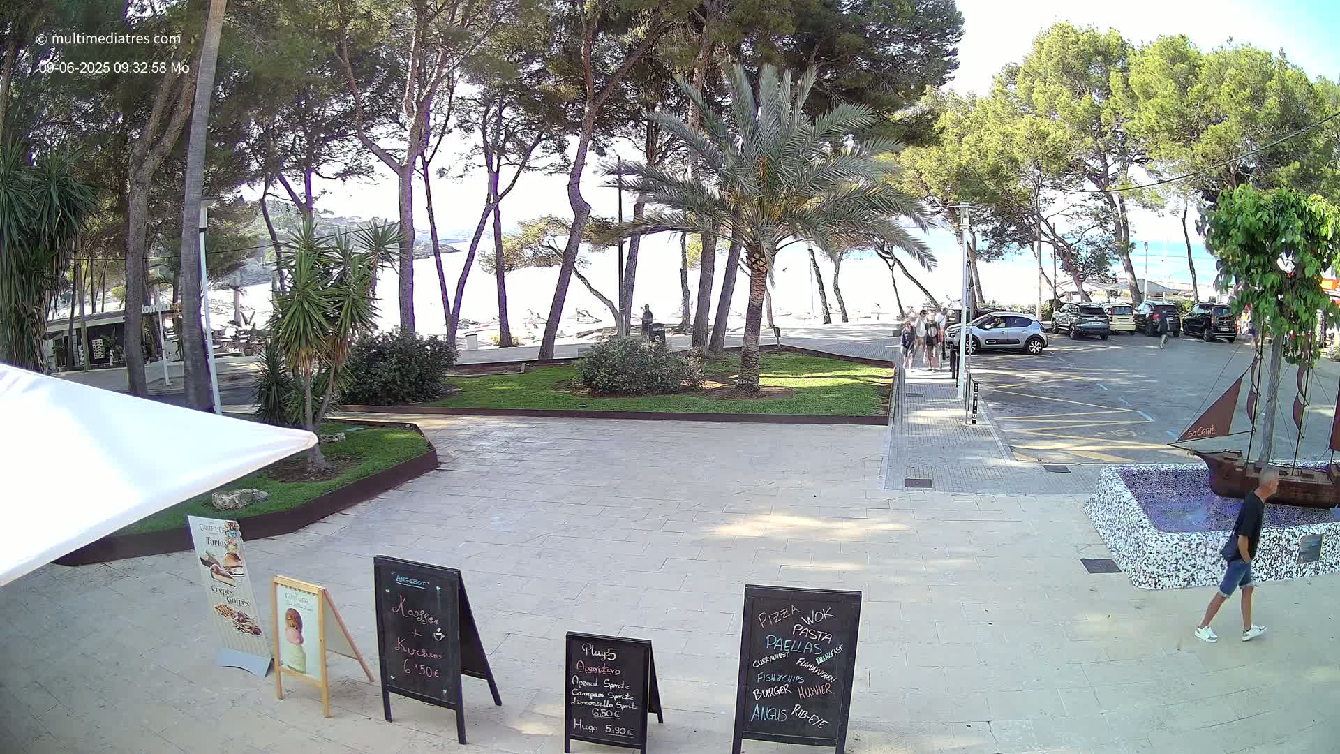 A paved plaza with palm trees and pine trees, overlooking a sandy beach on a sunny day, features several parked cars and a small boat-shaped fountain.