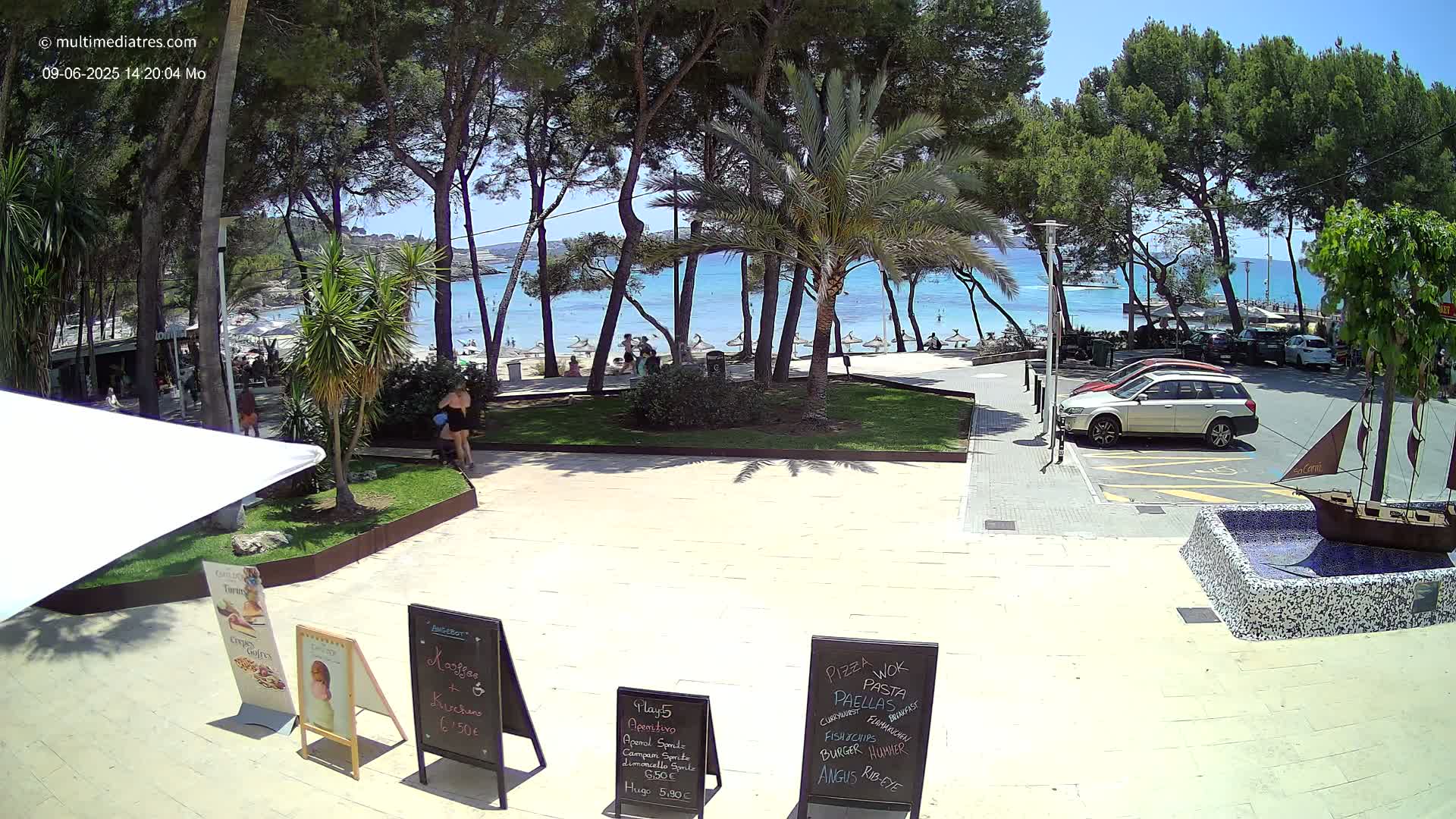 A sunny beachfront scene shows a paved area with parked cars, people on a sandy beach beyond trees, and several menu boards in front of an outdoor cafe.