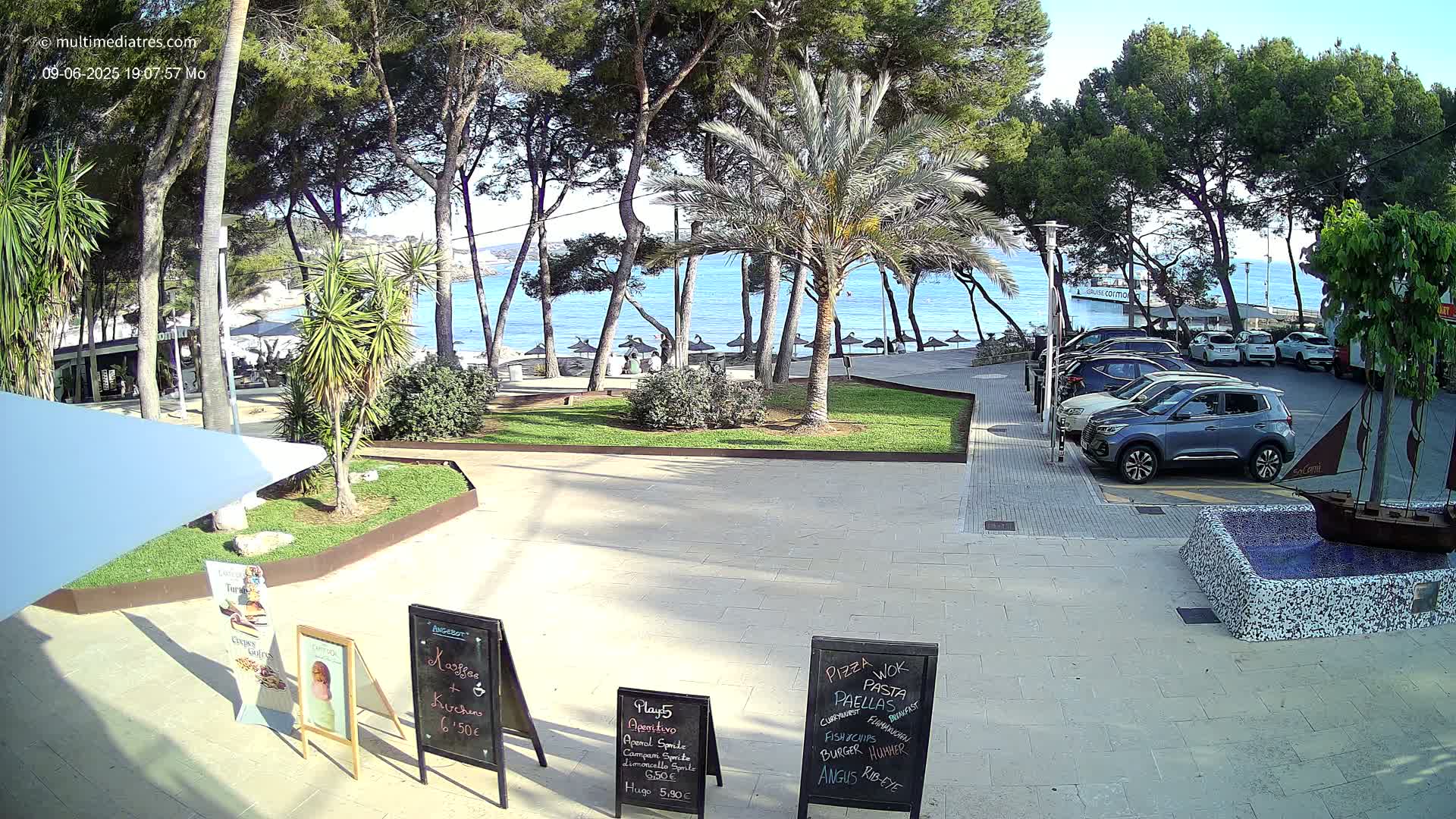 A paved plaza with palm trees and other vegetation overlooks a calm, blue ocean beach with umbrellas and a few people, under a sunny sky; several cars are parked nearby.