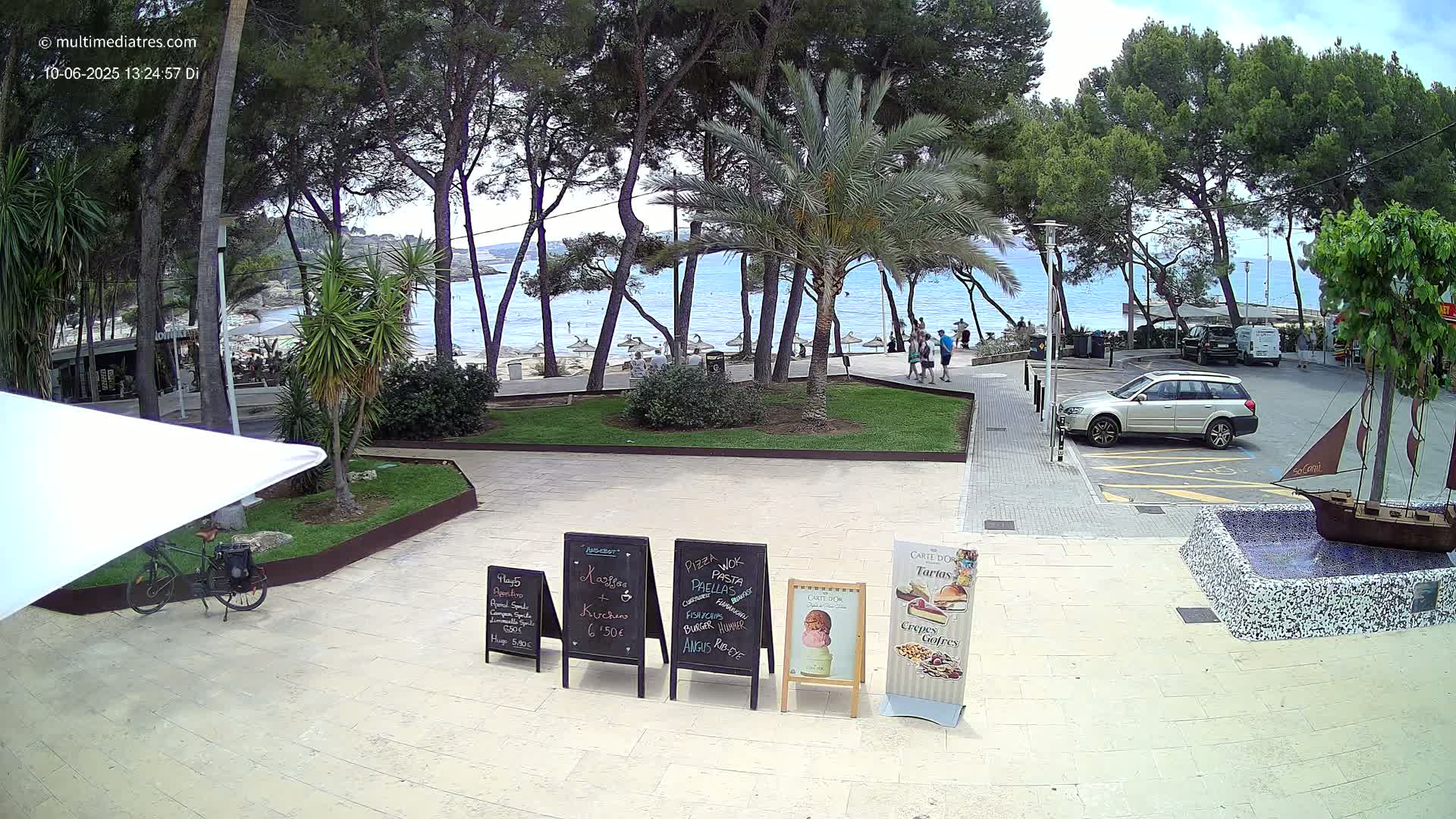 A partly sunny outdoor scene shows a paved area with several menu boards near a bicycle, a parked silver SUV, and a small model ship in a fountain, all set against a backdrop of trees, a beach, and calm blue water.