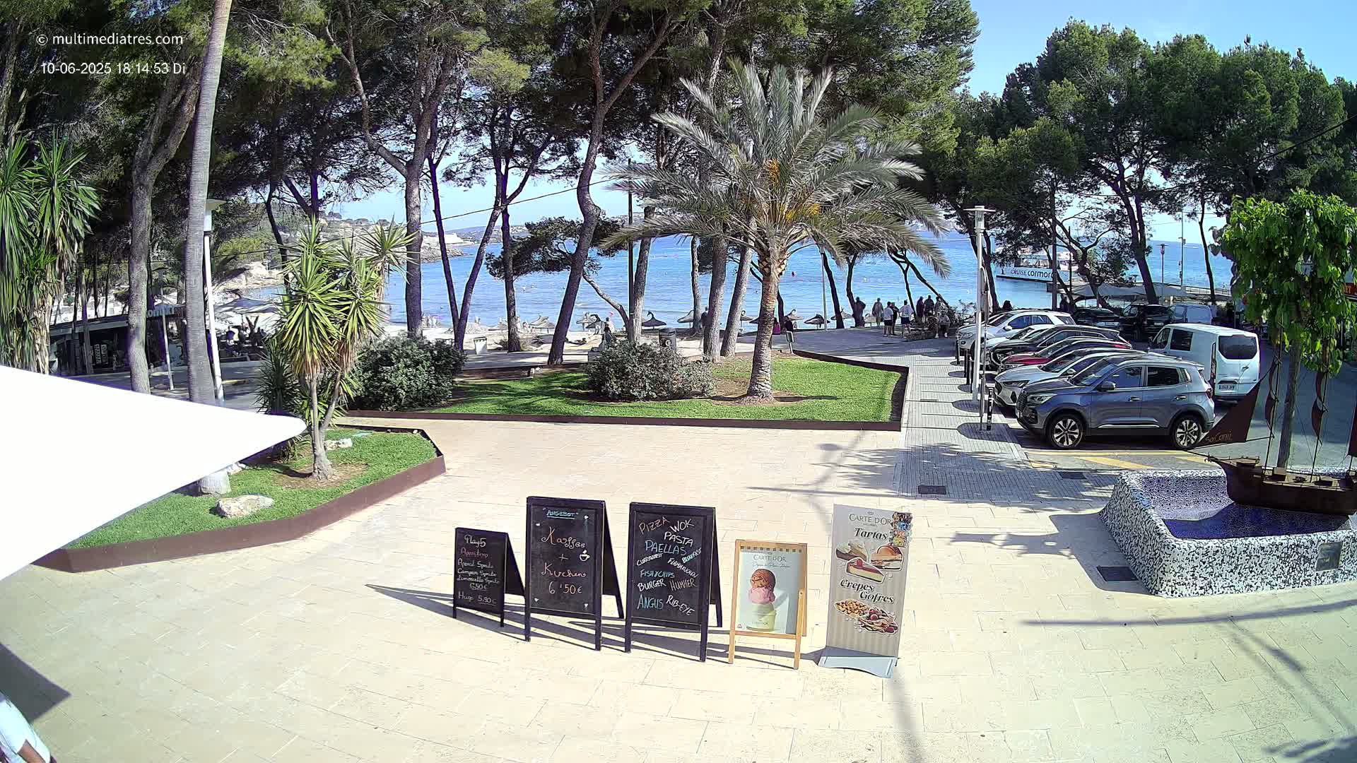 A sunny seaside plaza scene shows a calm ocean, a sandy beach, palm trees, parked cars, and several outdoor restaurant menu boards.