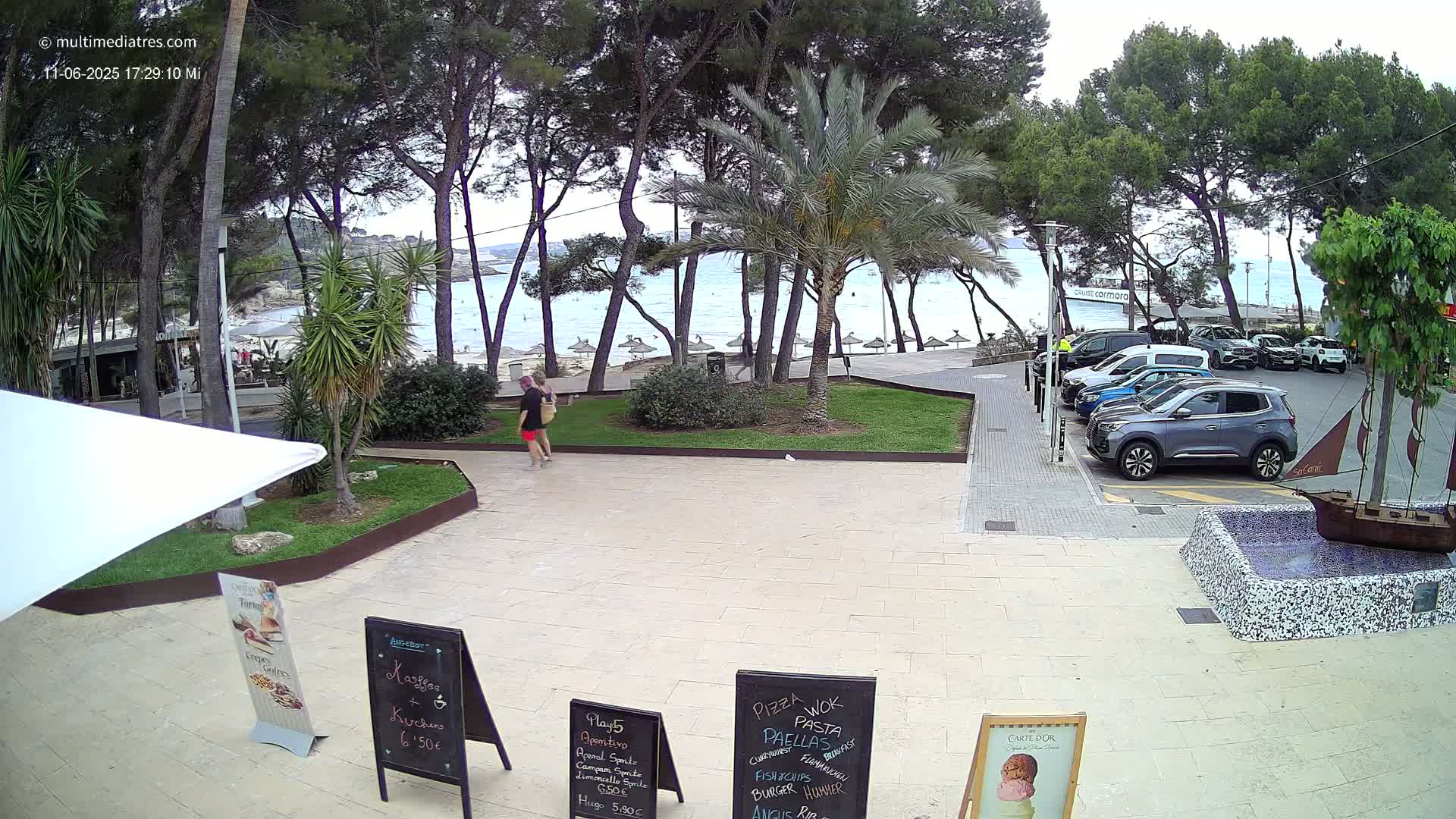 A partly cloudy day at a beachside restaurant with people walking, cars parked nearby, and palm trees swaying in a grassy area.