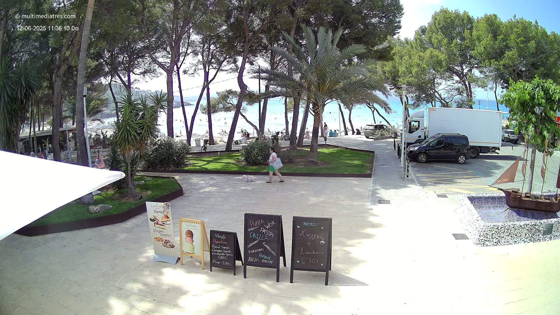 A woman walks a small white dog past outdoor restaurant menus near a sunny beach with people swimming and lounging, under a clear sky.