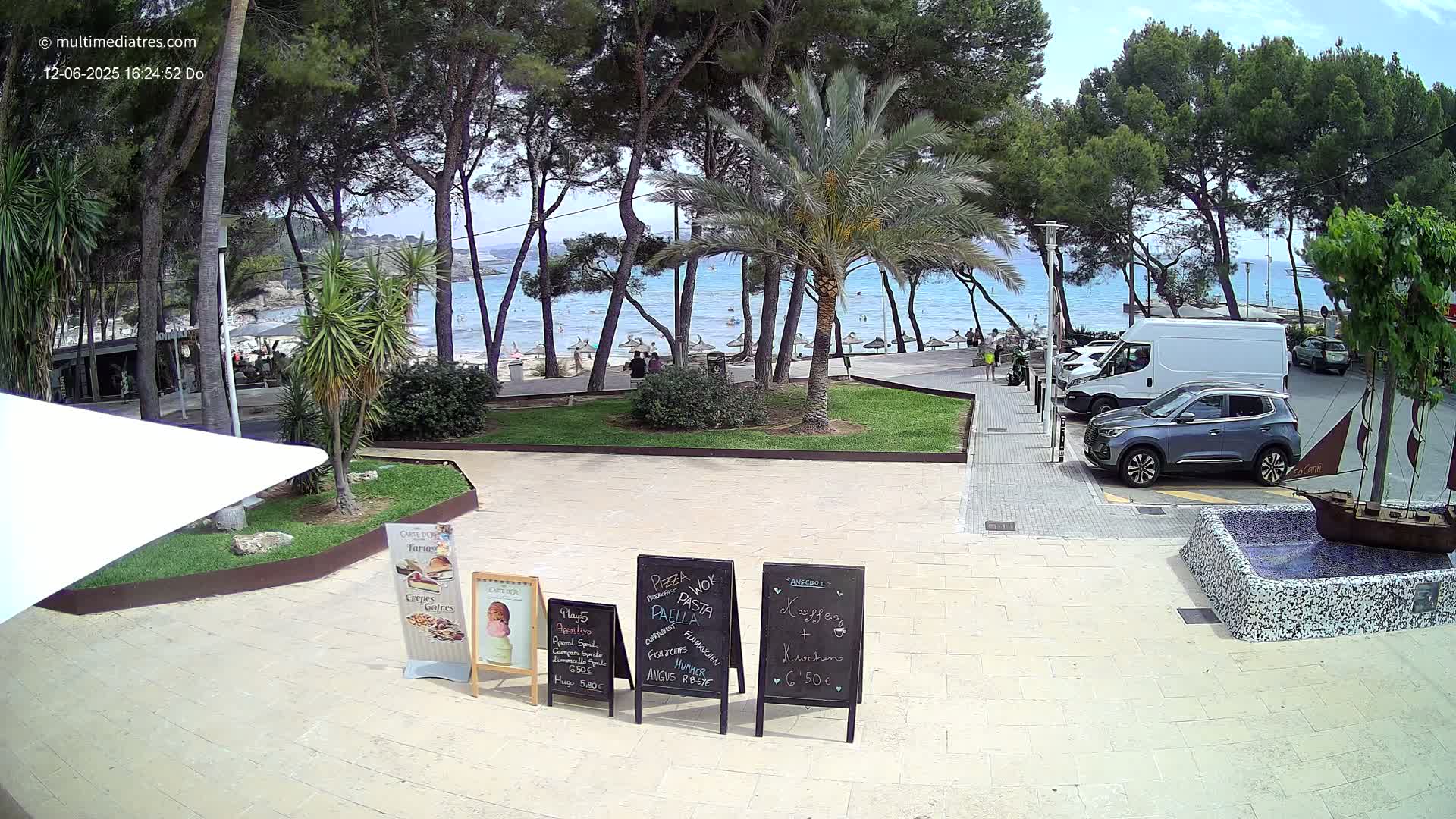 A sunny beachfront scene shows a paved plaza with several cars parked, palm trees, a grassy area, and a sandy beach with people swimming in a calm blue ocean.