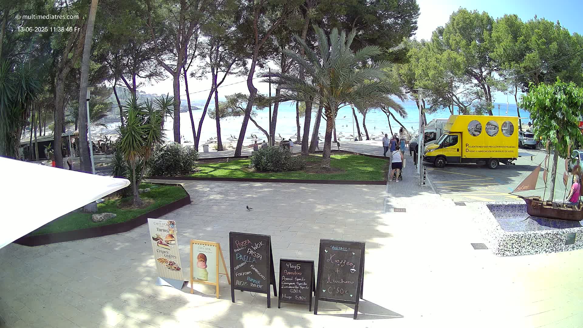 A sunny beachfront scene shows a paved area with several menu boards, a small decorative ship, and a yellow delivery truck parked near a walkway leading to a sandy beach with people relaxing under a bright sky.