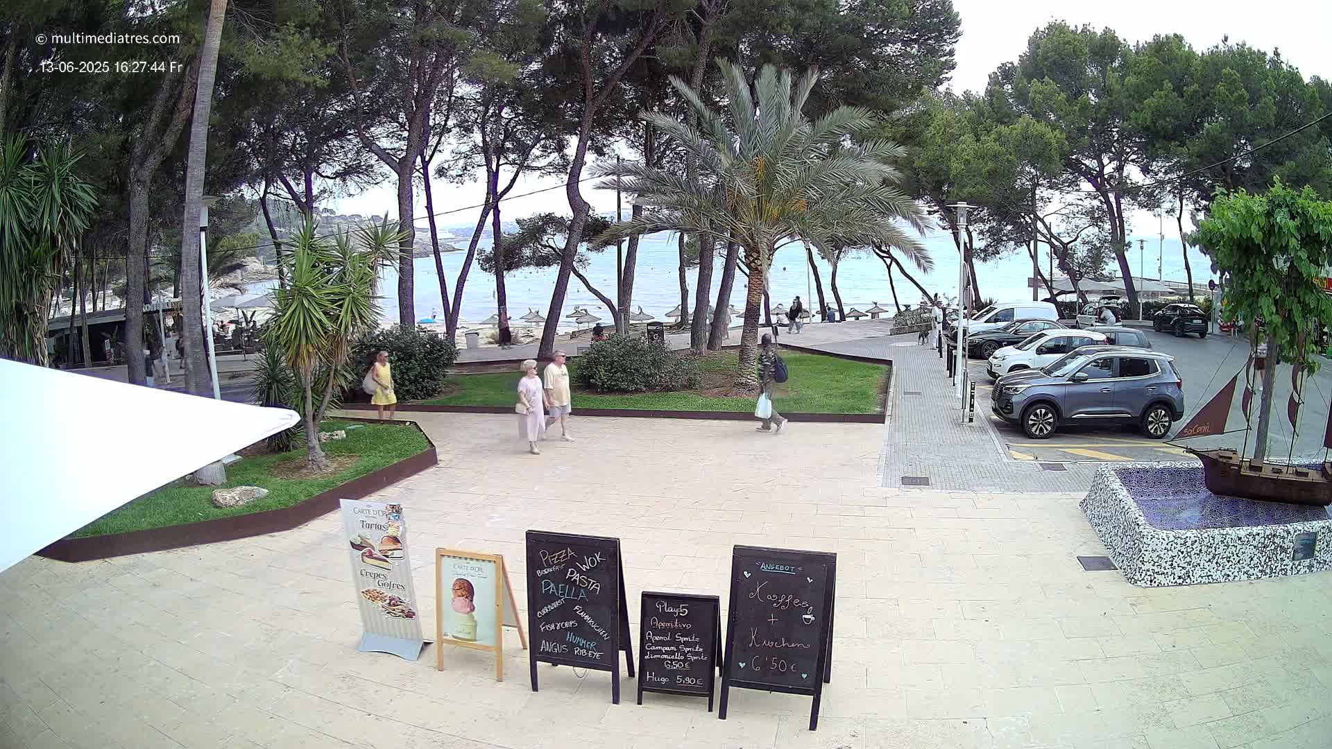 A seaside patio scene under an overcast sky shows several people walking past palm trees and parked cars toward a sandy beach.