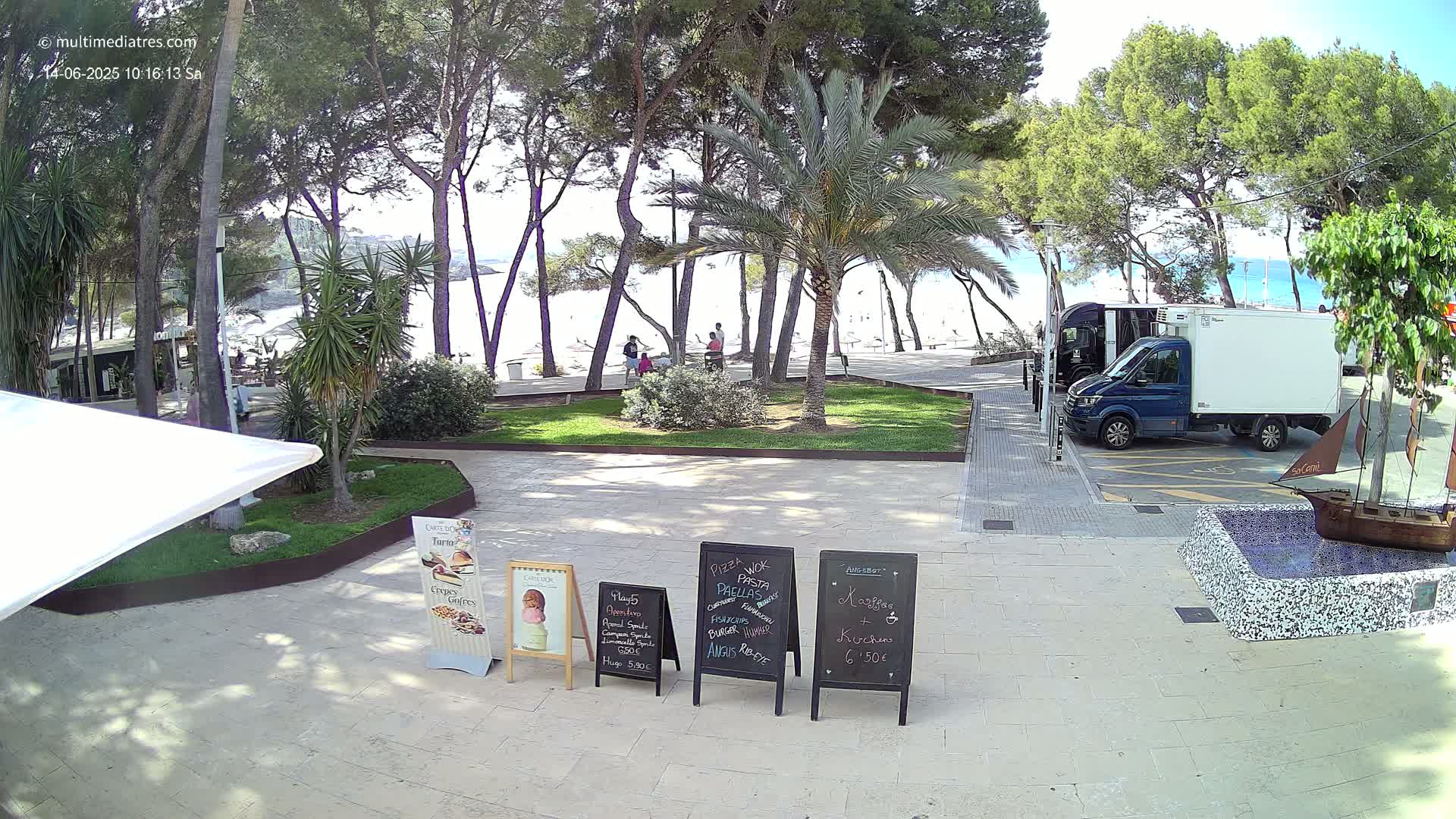 A sunny outdoor scene shows a paved area with several menu boards, a partially visible white canopy, a small decorative fountain, and a blue delivery van parked near a beach visible through trees.