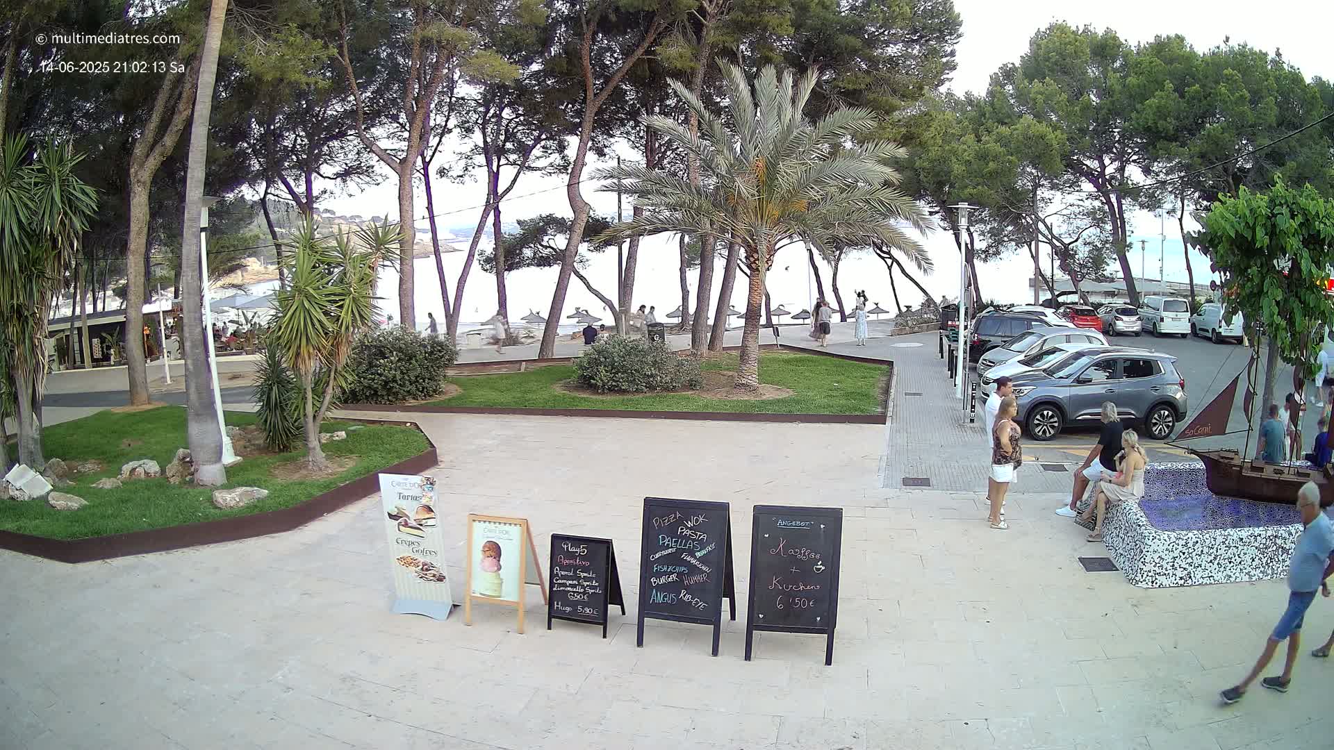 A sunny beachfront scene shows a parking lot near a beach with people swimming and relaxing,  palm trees and other vegetation, and several menu boards.