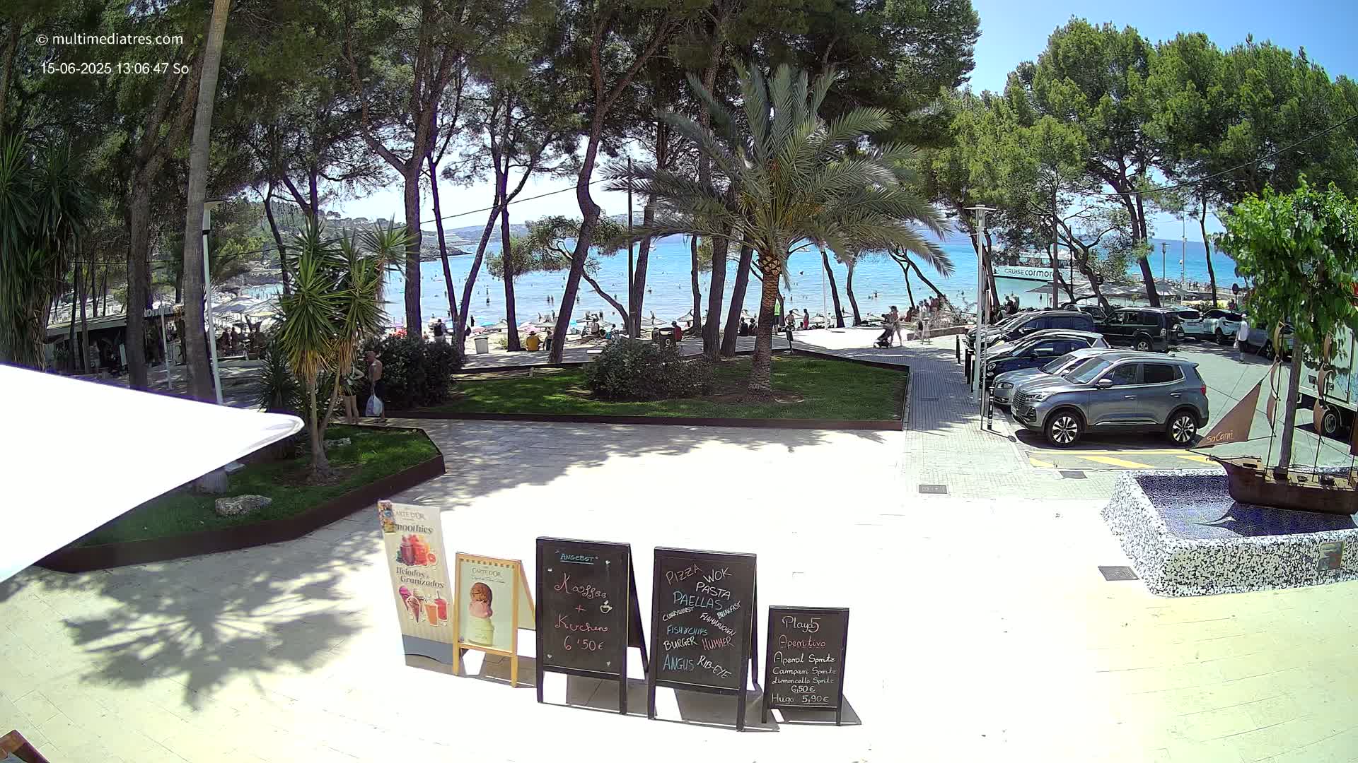 A sunny beach scene is viewed from a patio area with palm trees, parked cars, and several menu boards showing food and drink options.