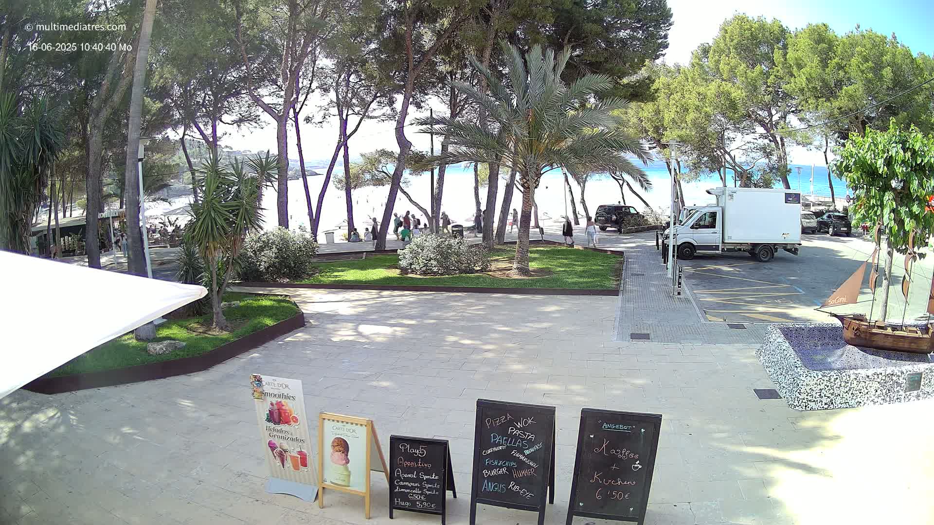 A sunny outdoor scene shows a paved area with palm trees and other vegetation, a sandy beach and ocean in the background, and several people walking along a path near the beach.