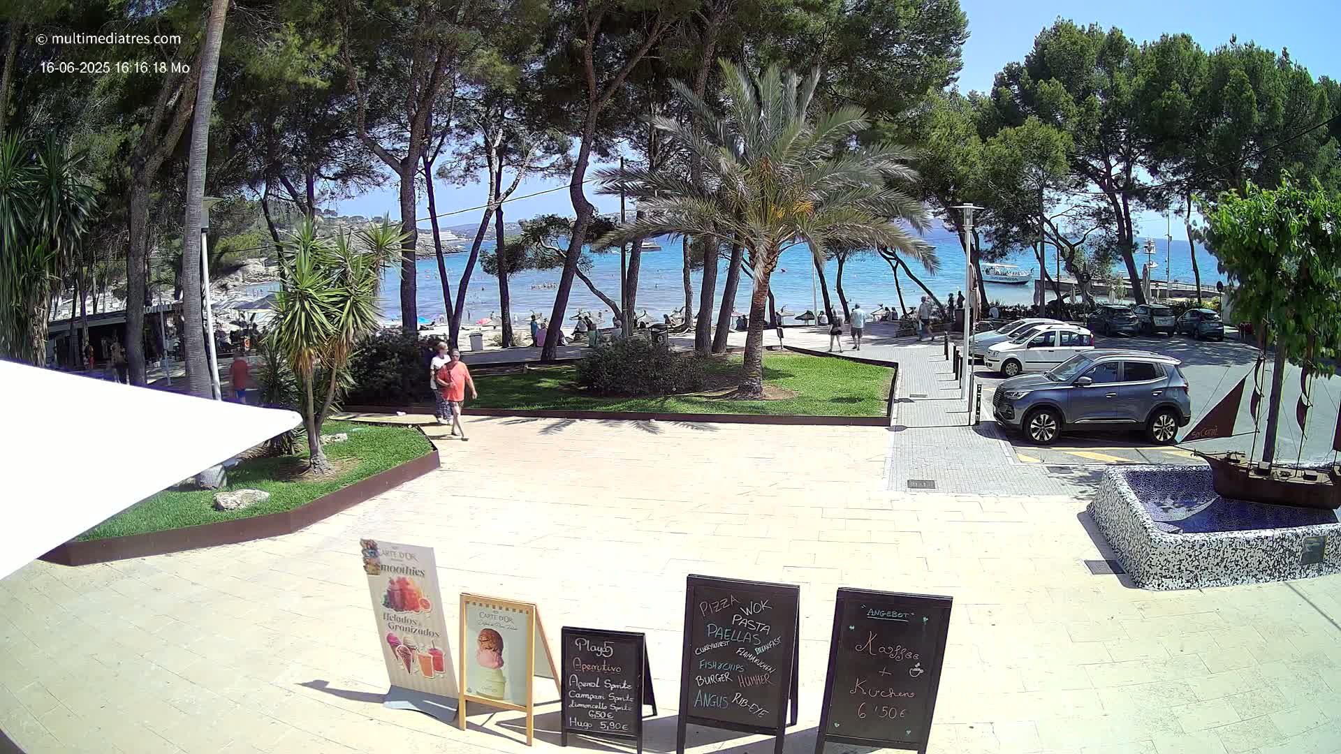 A sunny beach scene with turquoise water, palm trees, and parked cars is viewed from a patio area with outdoor menu boards.
