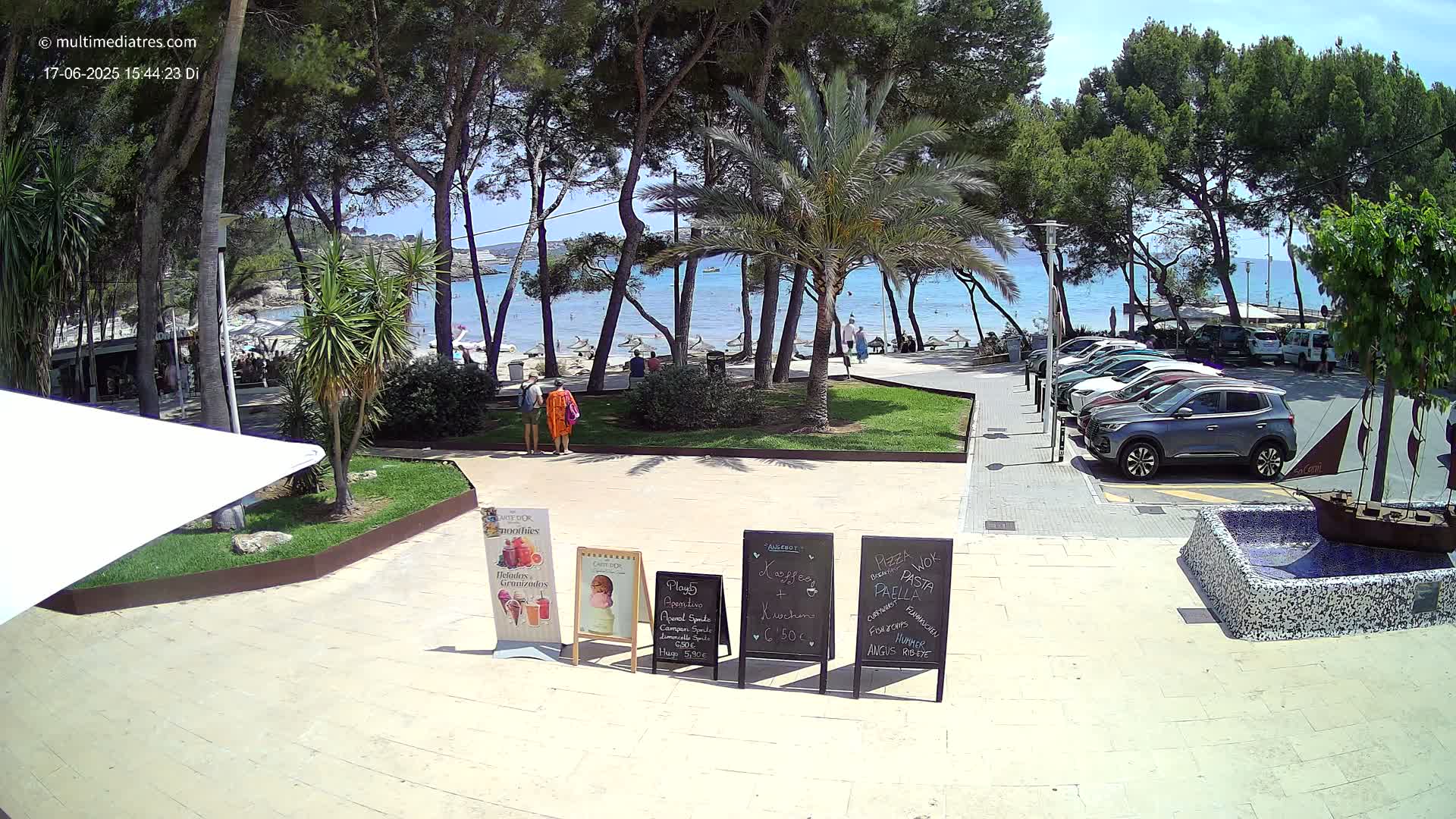 A sunny outdoor scene shows a paved area with several menu boards, a small fountain, and a parking lot next to a beach with people swimming in the ocean under a clear blue sky.