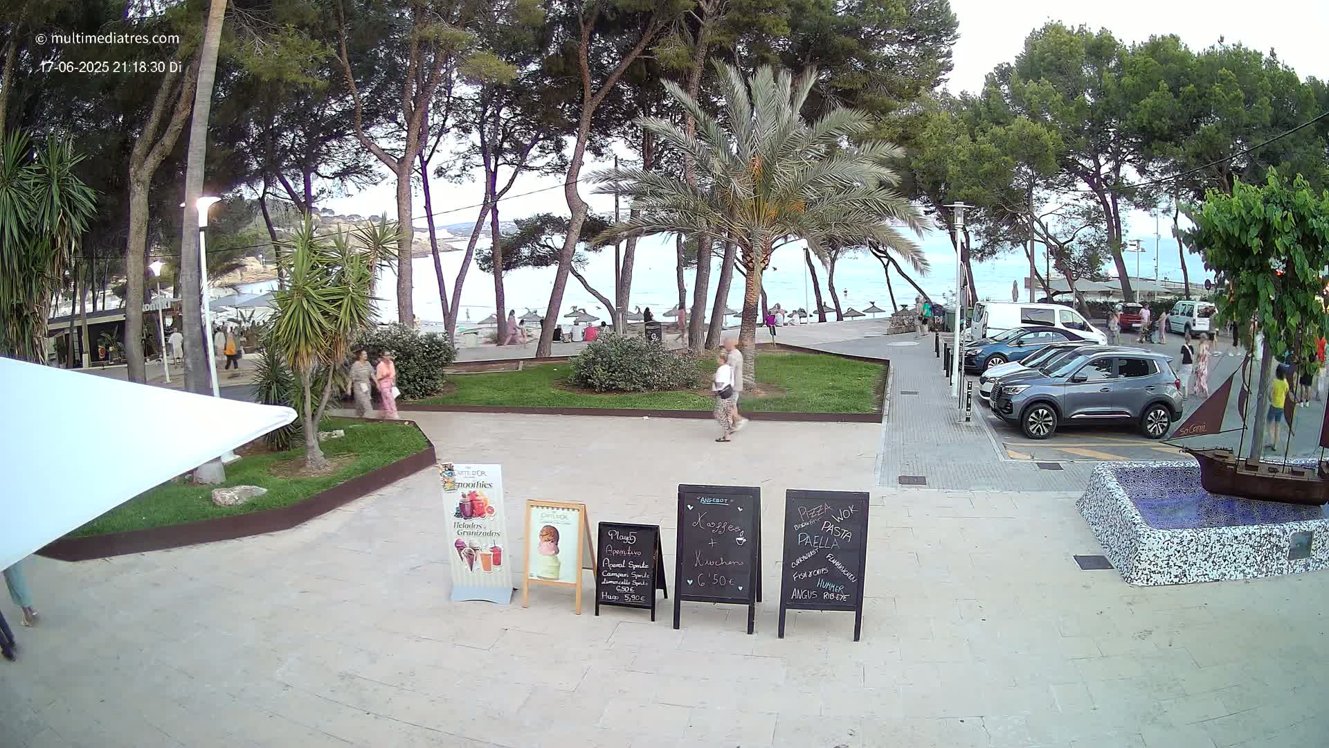 A paved seaside plaza with parked cars, people strolling, and several menu boards, under a partly cloudy evening sky.