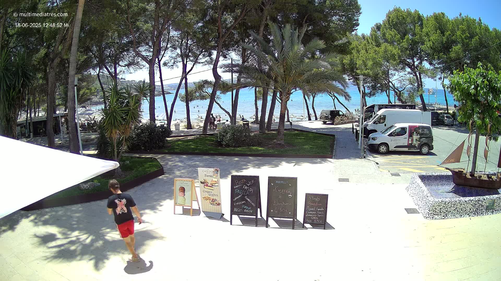 A sunny beach scene is visible through a line of trees bordering a paved area with several menu boards and a person walking, with several parked vans and a small decorative boat fountain in the foreground.