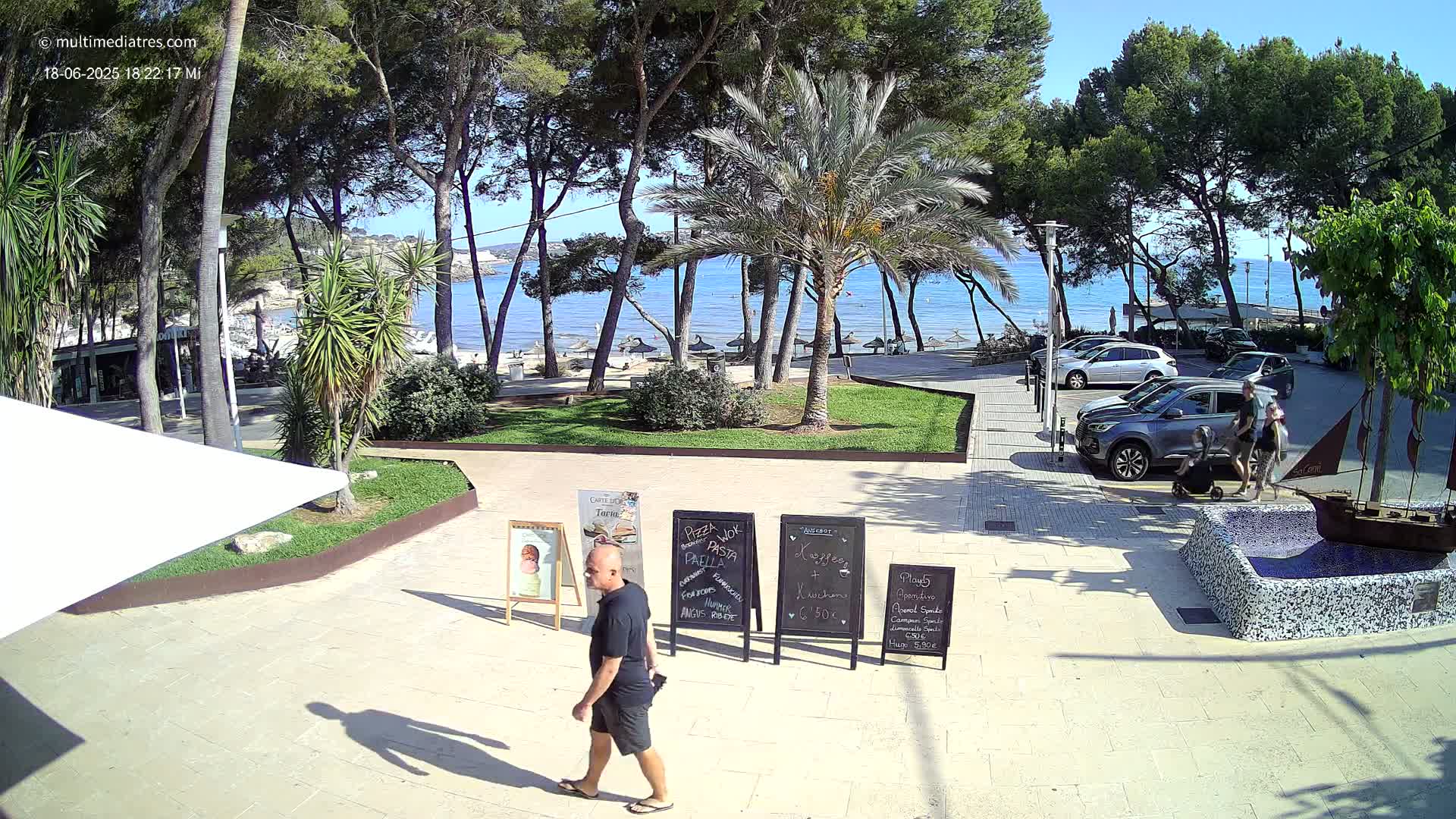 A man walks past outdoor restaurant menus toward a beach, which is partially visible beyond a parking lot and a row of trees under a sunny sky.