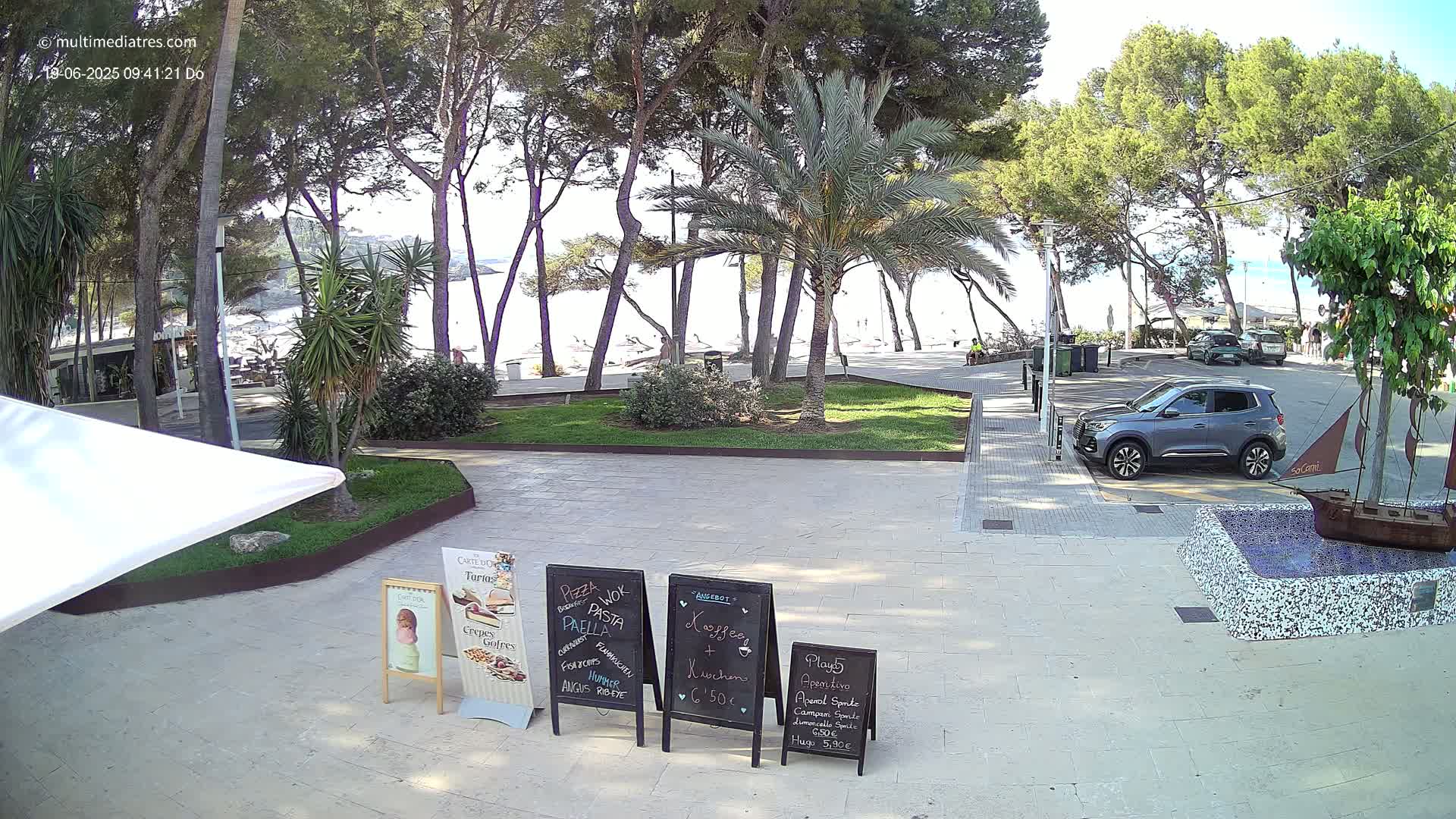 A paved area with several palm trees and other greenery, a beach visible in the background under a clear sky, and a gray SUV parked nearby.