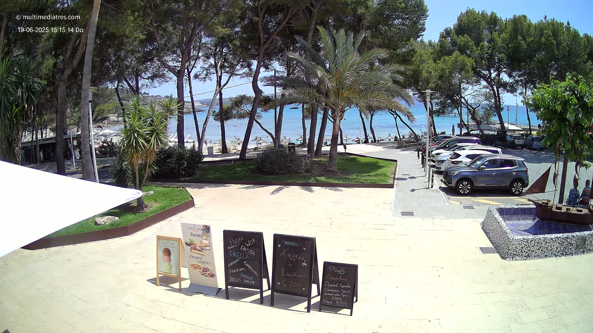 A sunny outdoor scene shows a parking lot with several cars near a restaurant with menu boards, overlooking a beach with people and calm blue water beyond a line of trees.