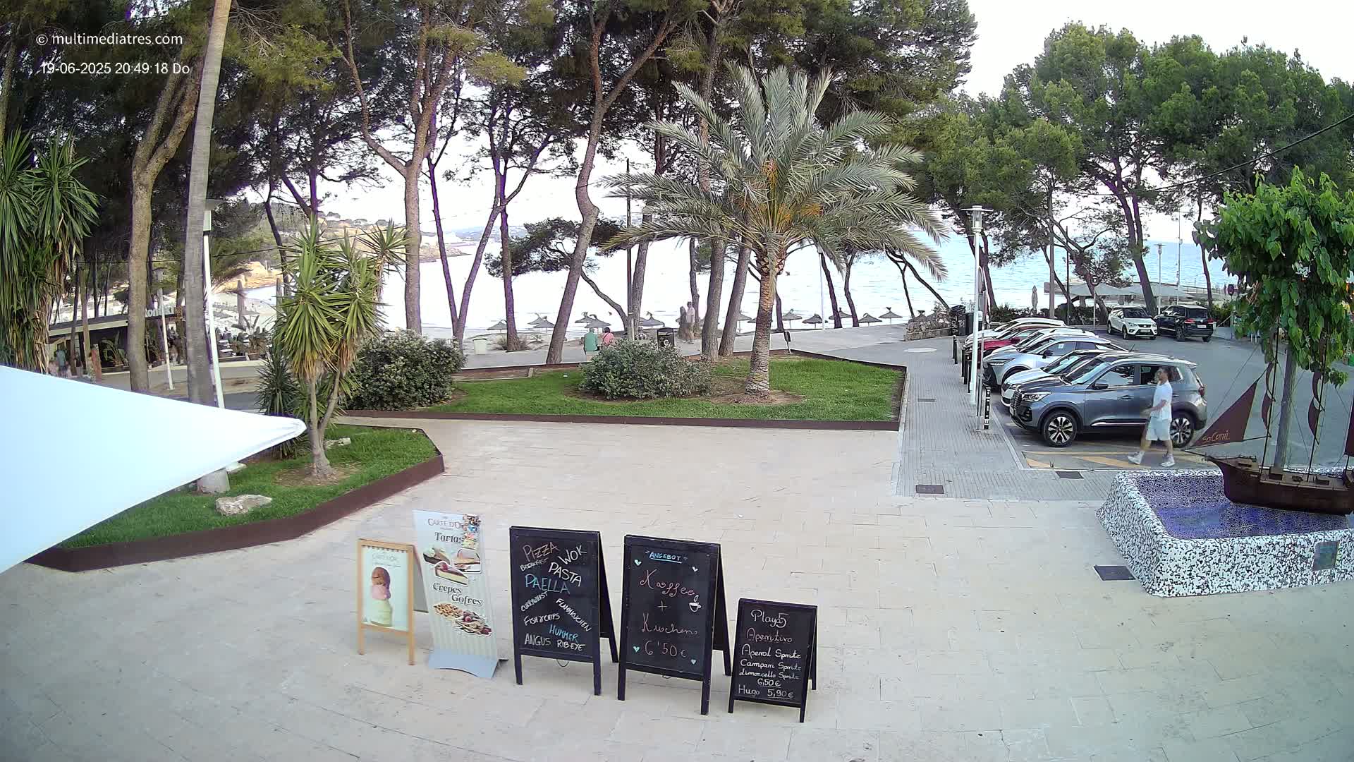 A paved plaza with several menu boards, a small fountain, and a few parked cars, overlooks a beach with calm water and many trees under a partly cloudy sky.